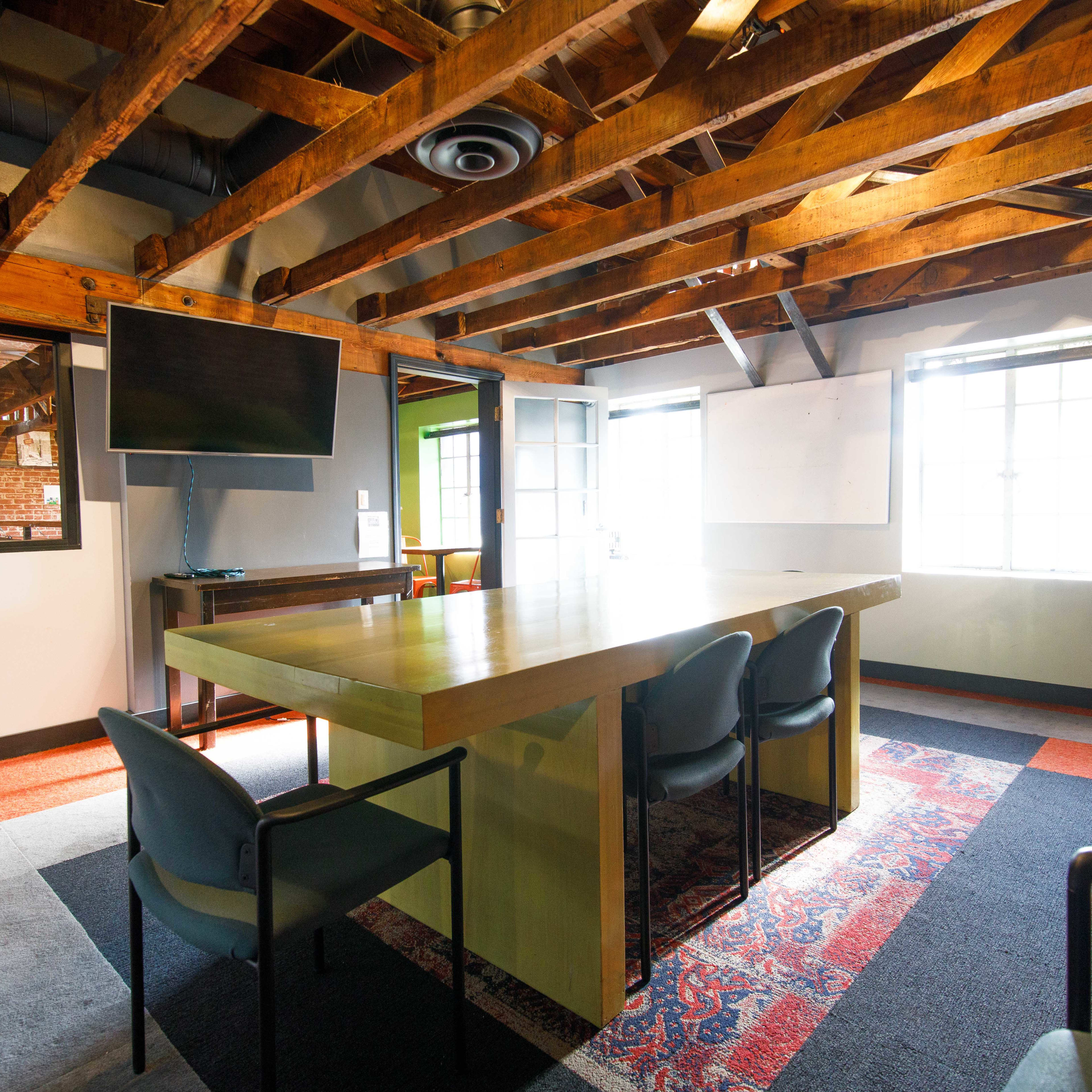 A modern meeting room featuring a long table, chairs, a television mounted on the wall, and exposed wooden beams in the ceiling.