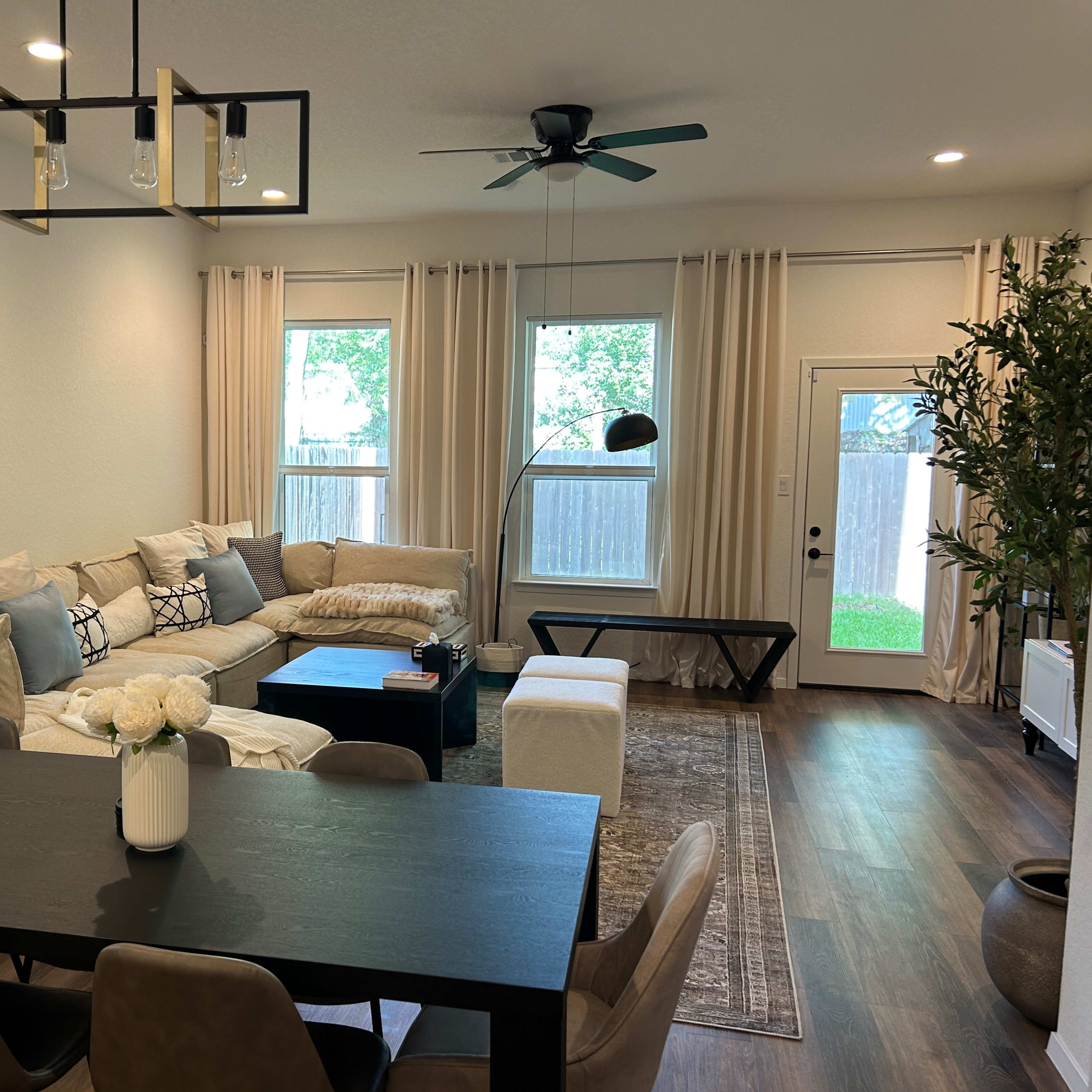 A living room with a sectional sofa, a dining table with chairs, and large windows providing natural light.