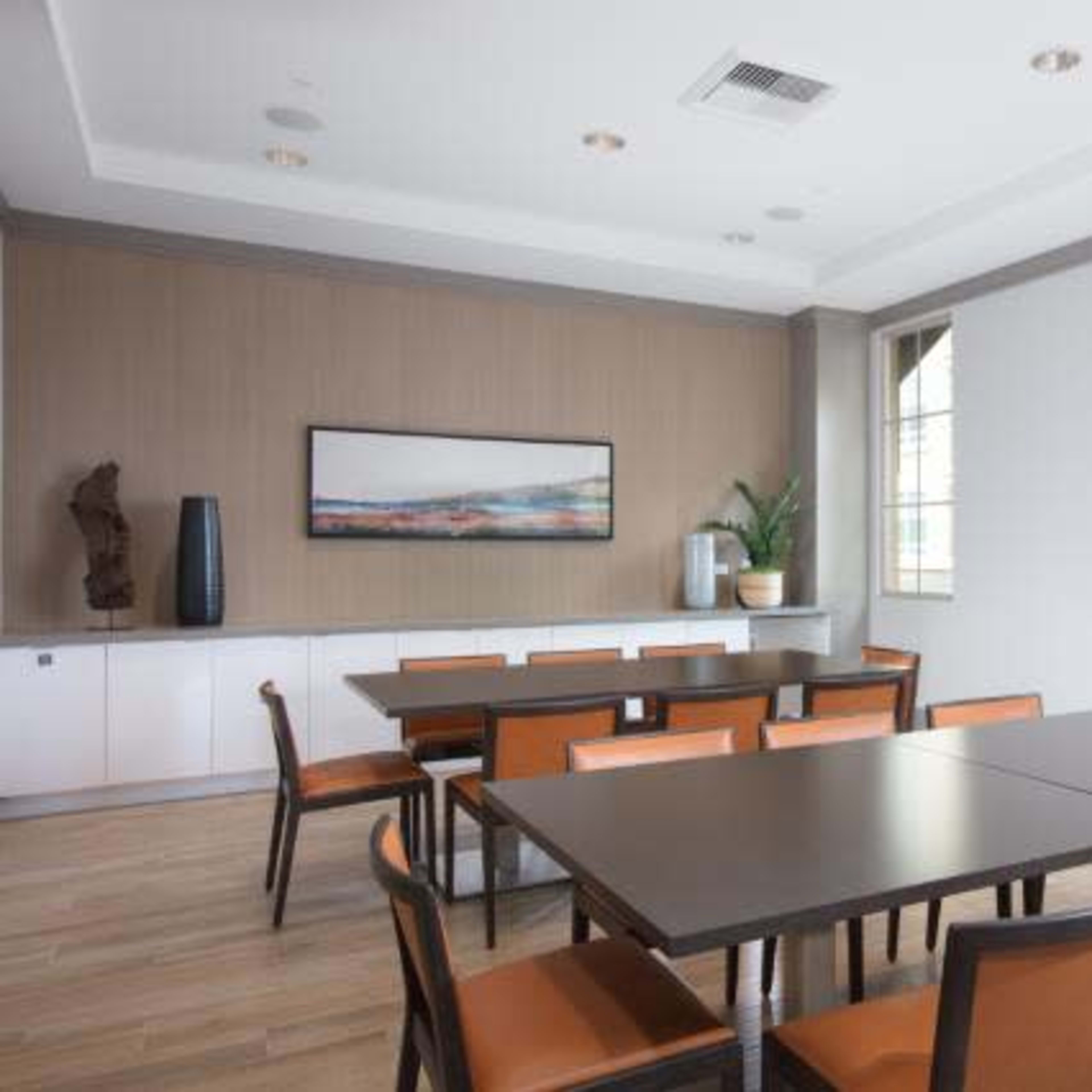 A meeting room features a large table surrounded by chairs, with a wall-mounted artwork and a plant near a window.