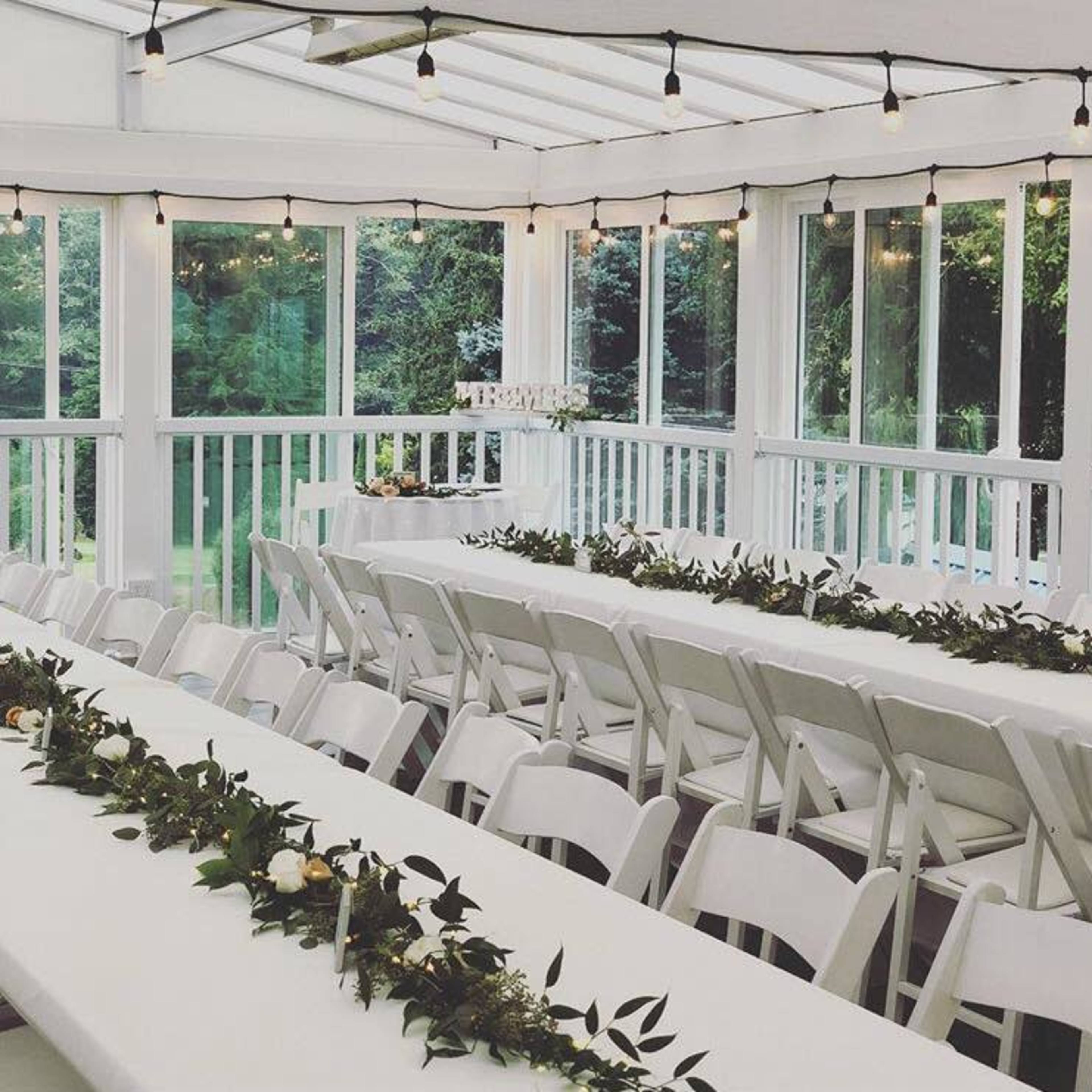 A bright room with white tables set for an event, featuring greenery centerpieces and string lights overhead.