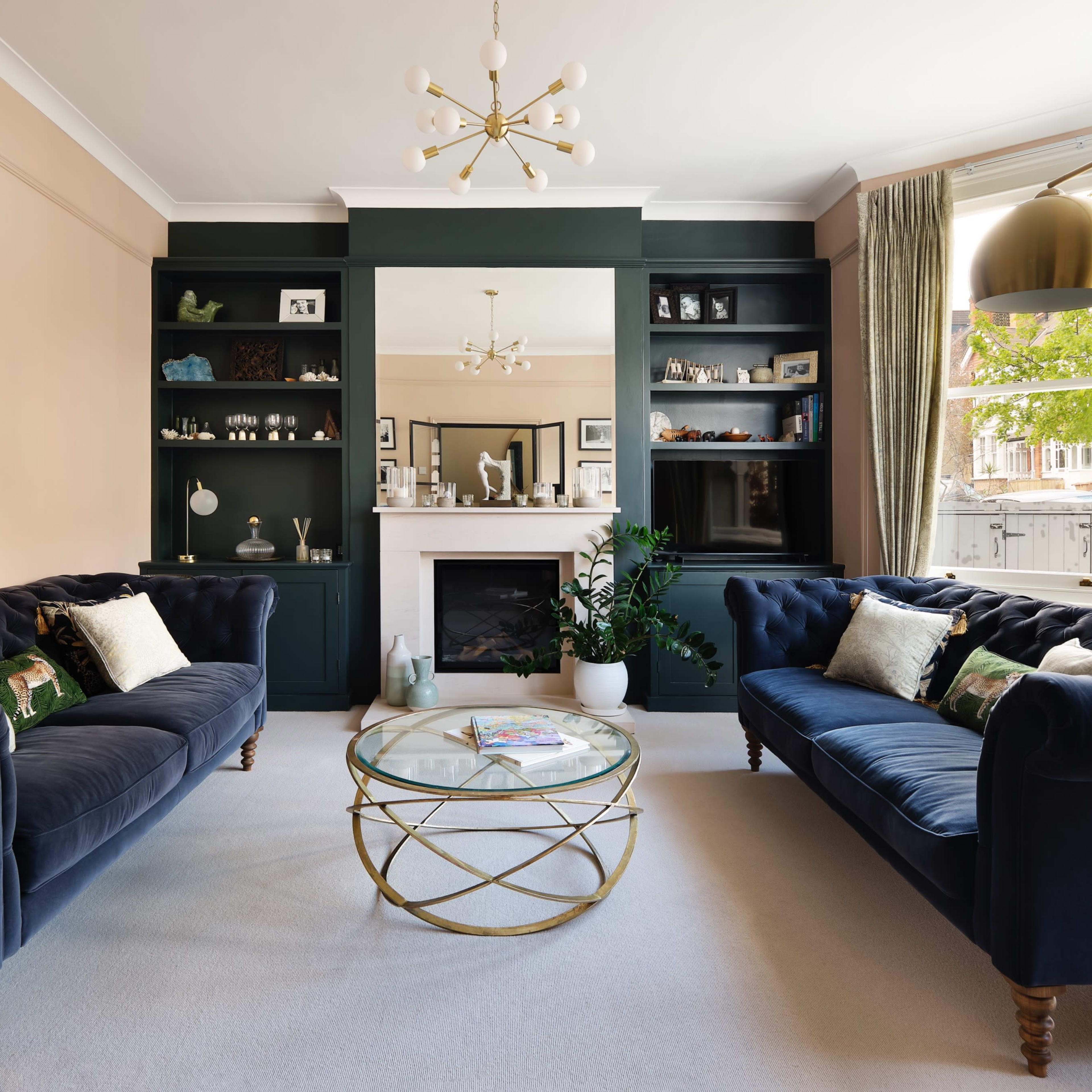 The living room features two blue velvet sofas facing a glass coffee table, with a modern chandelier and a fireplace surrounded by built-in shelves.