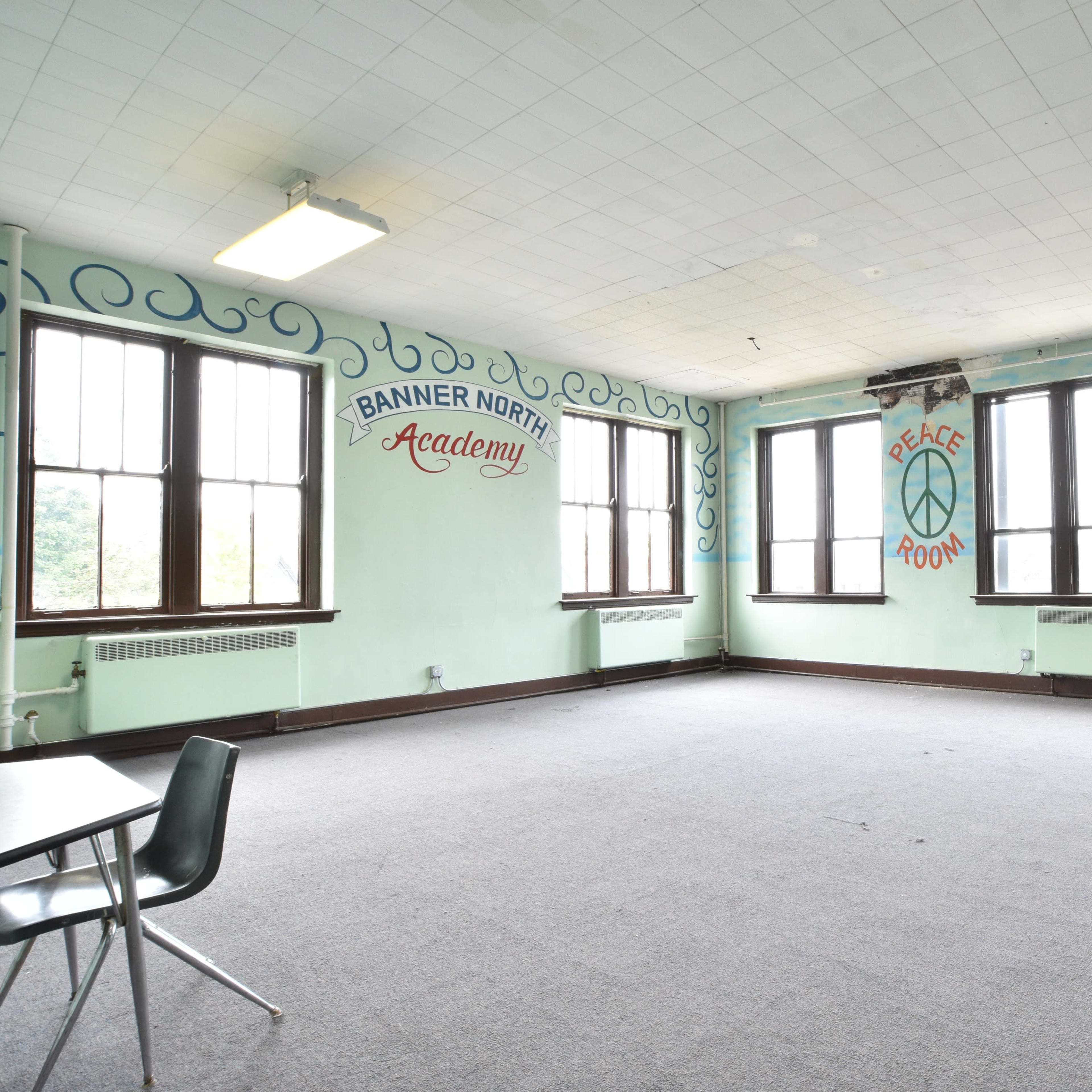 An empty classroom with large windows, light green walls featuring murals, and several tables and chairs arranged in the room.