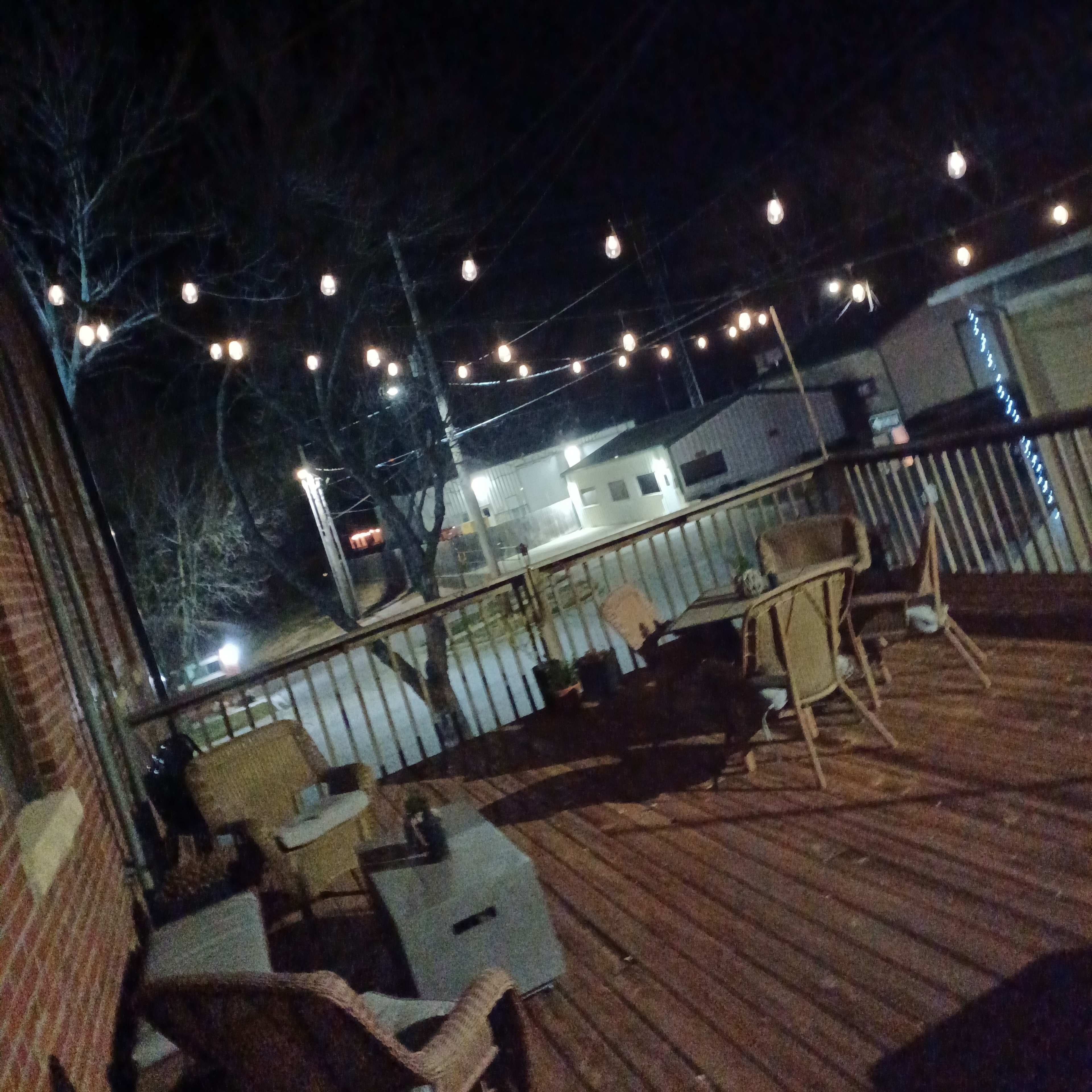 A wooden deck at night is illuminated by string lights, with several chairs arranged on it.