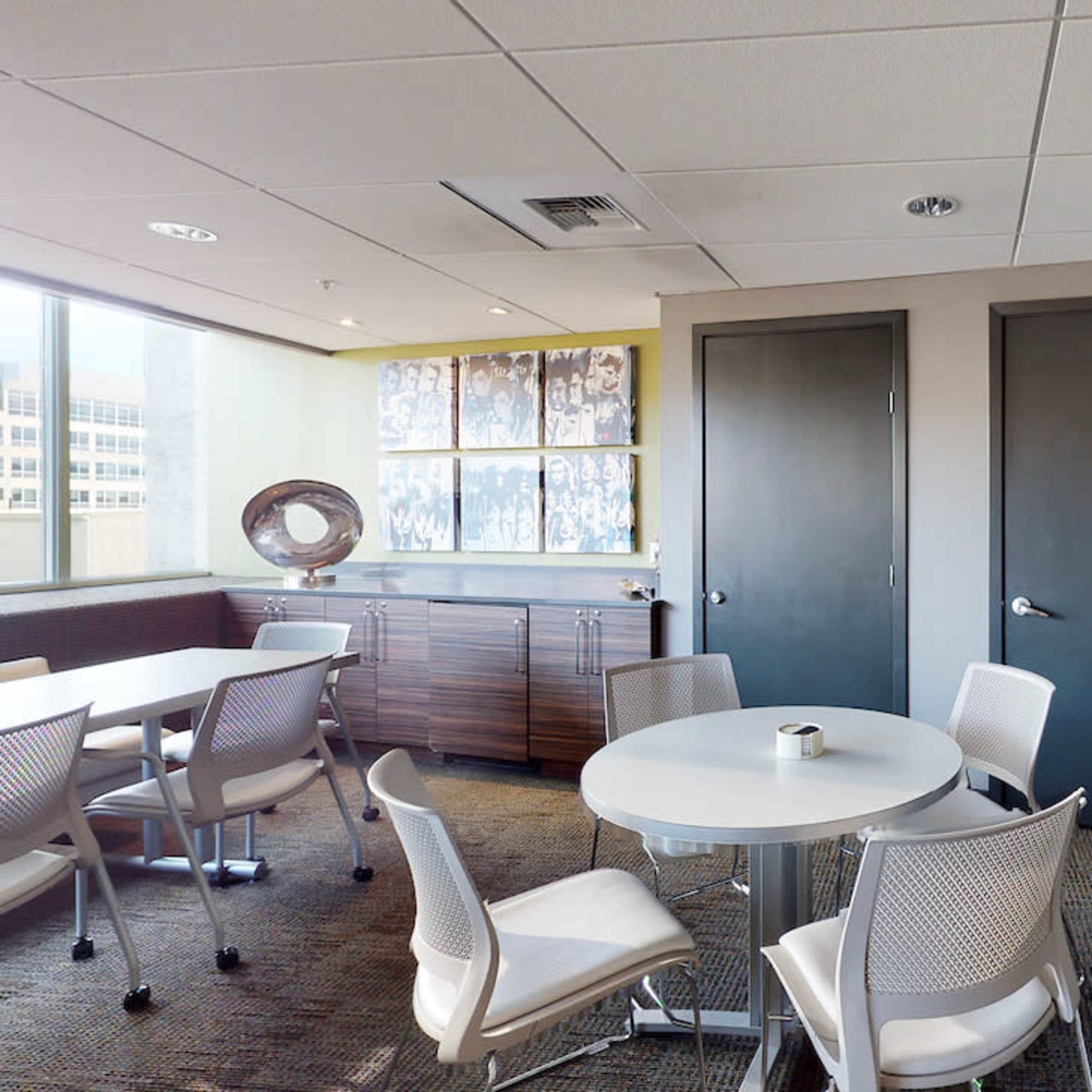 A modern office breakroom with a round table, chairs, a side cabinet, and two closed doors.