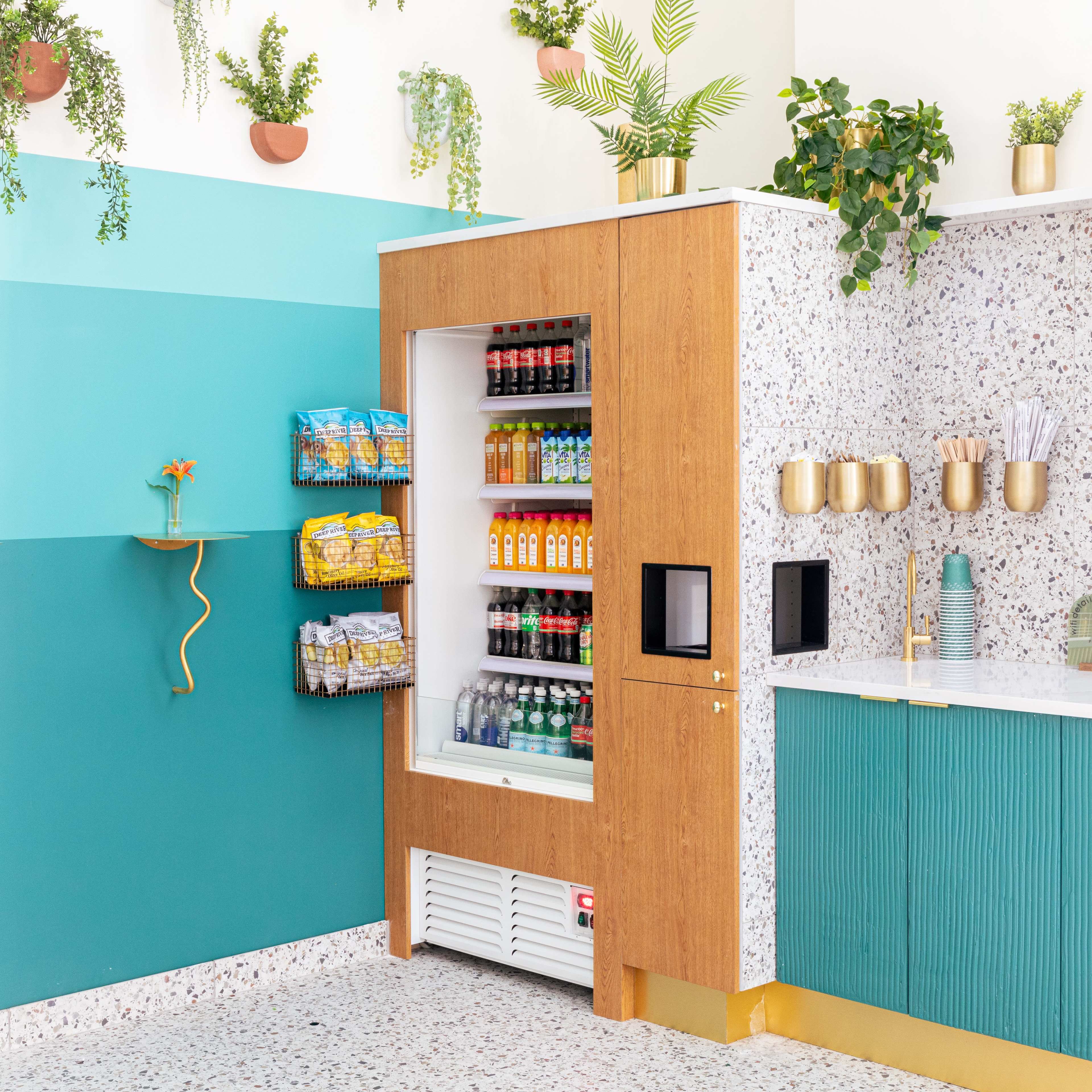 A modern snack and beverage station with a vending machine, shelving units, and a countertop adorned with plants and colorful items.