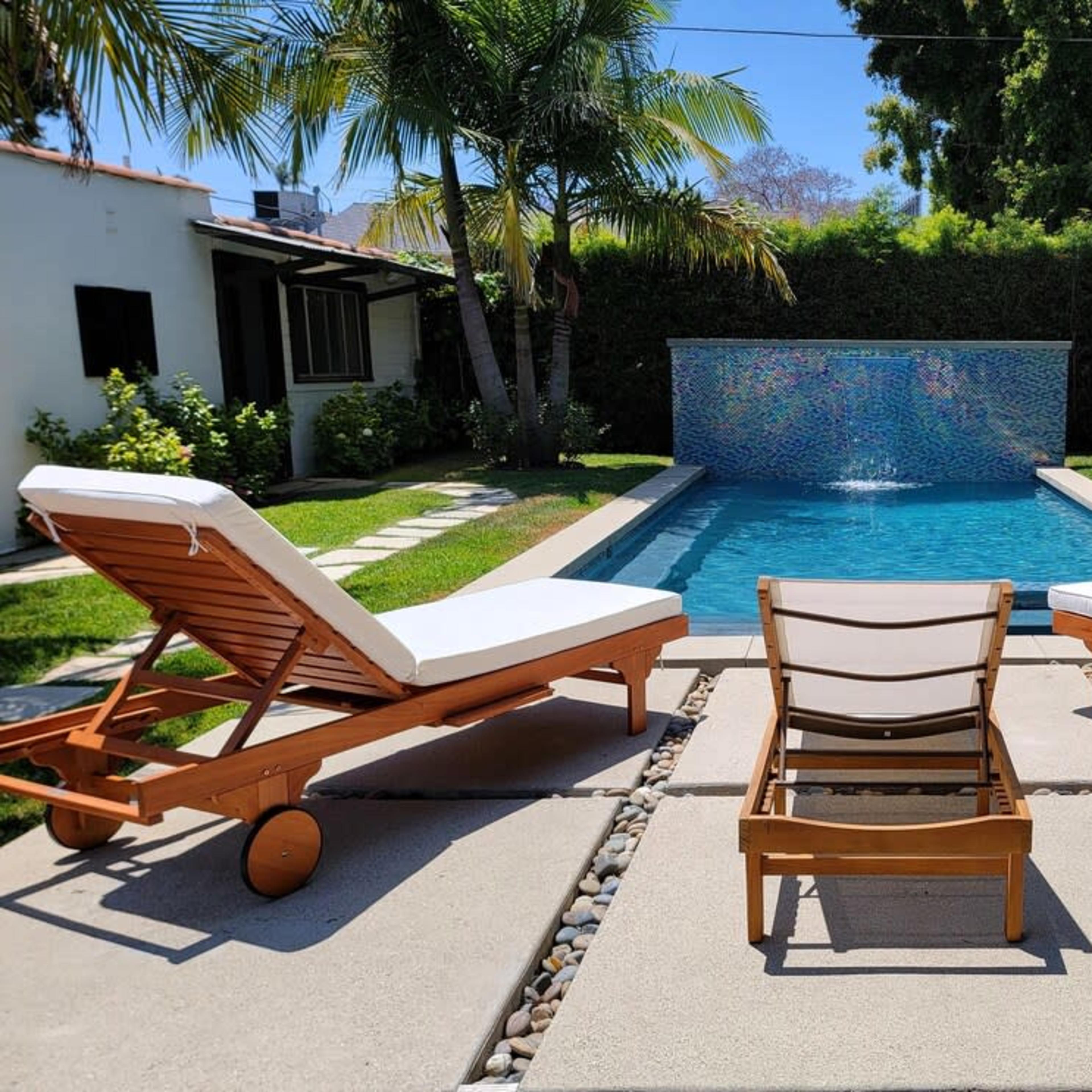 A serene outdoor area with two wooden lounge chairs beside a swimming pool, featuring a mosaic wall and lush greenery.