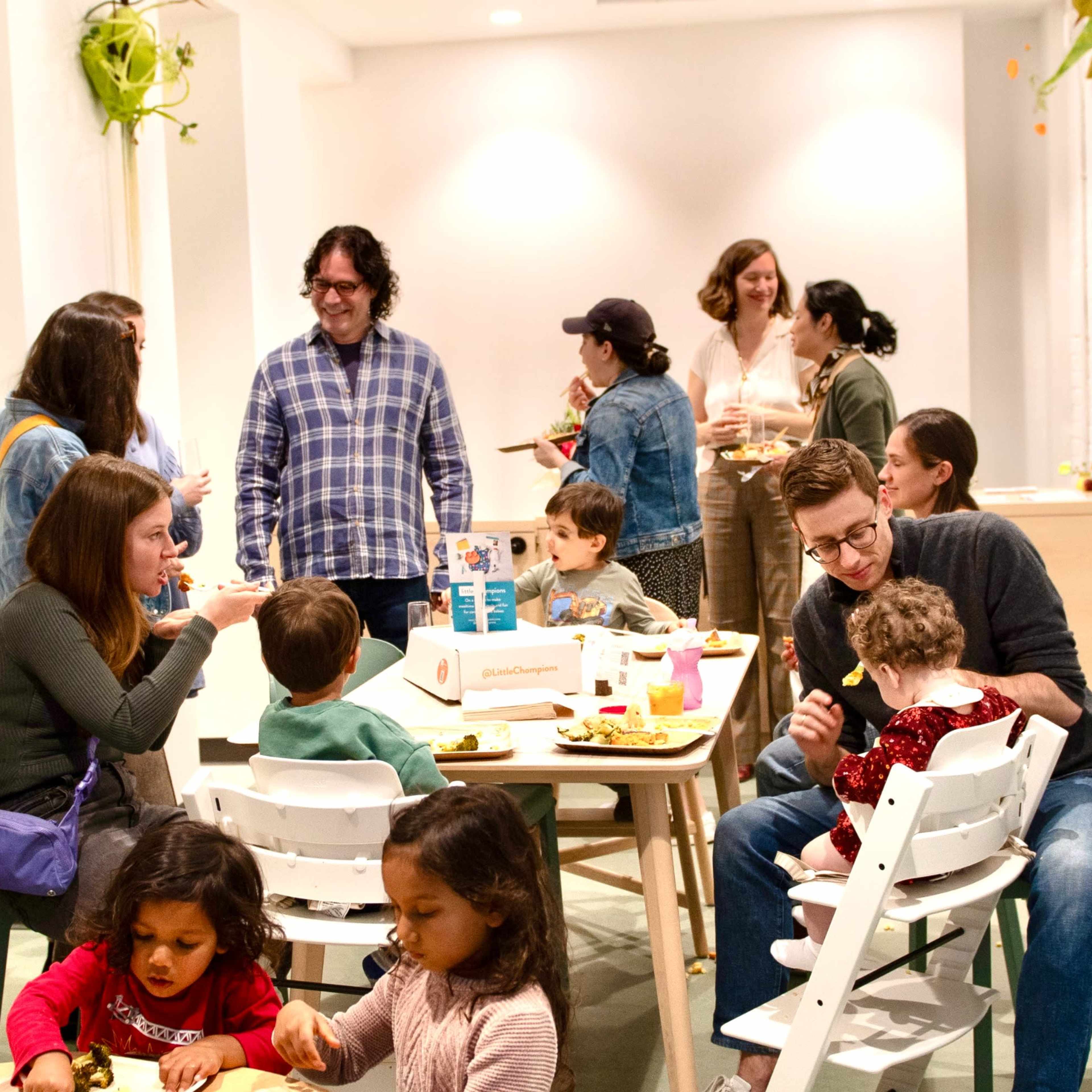 A group of adults and children share a meal and socialize in a brightly-lit room with a mix of seating arrangements and food containers on the tables.