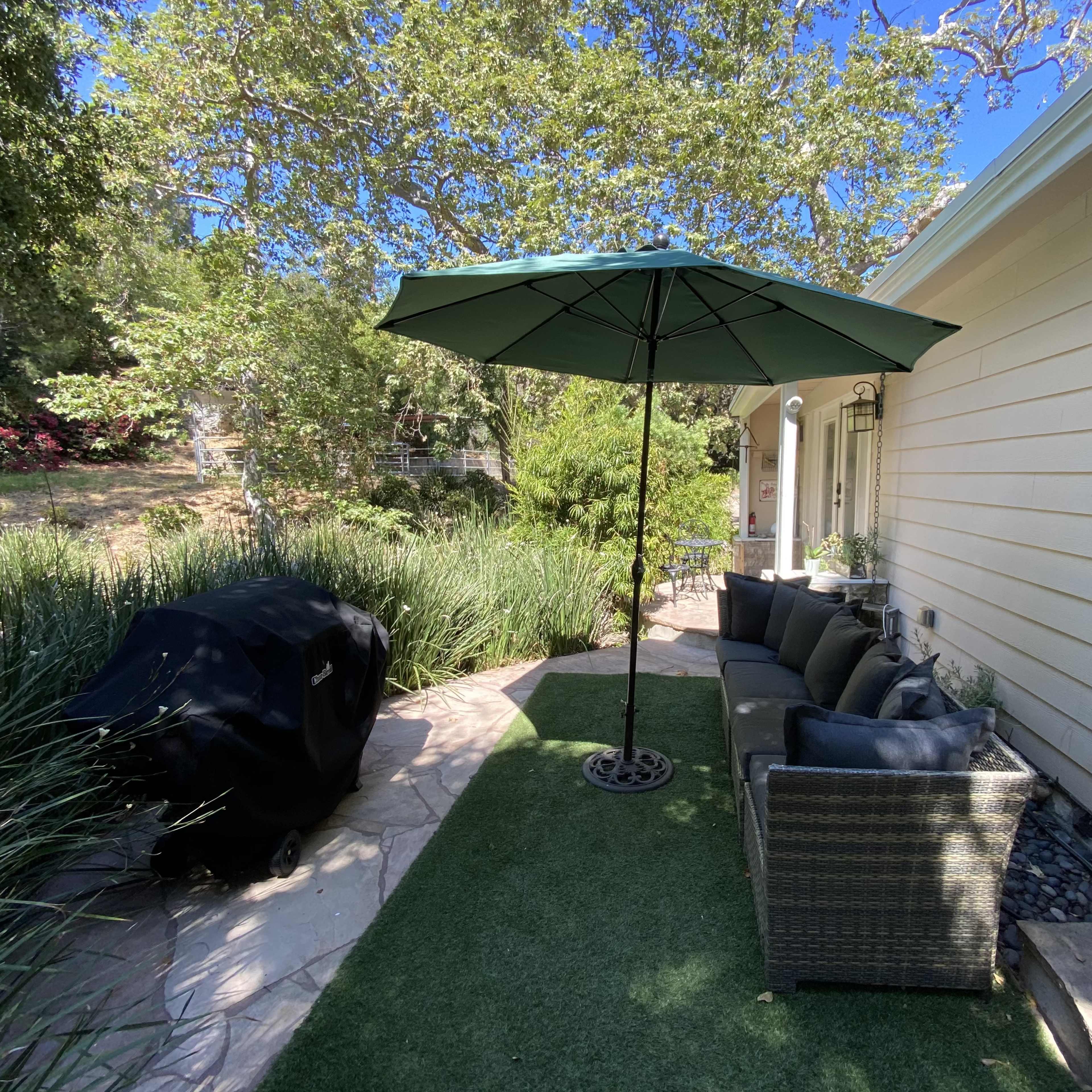 A patio area features a green umbrella over a seating arrangement and a grill beside a neatly landscaped garden.