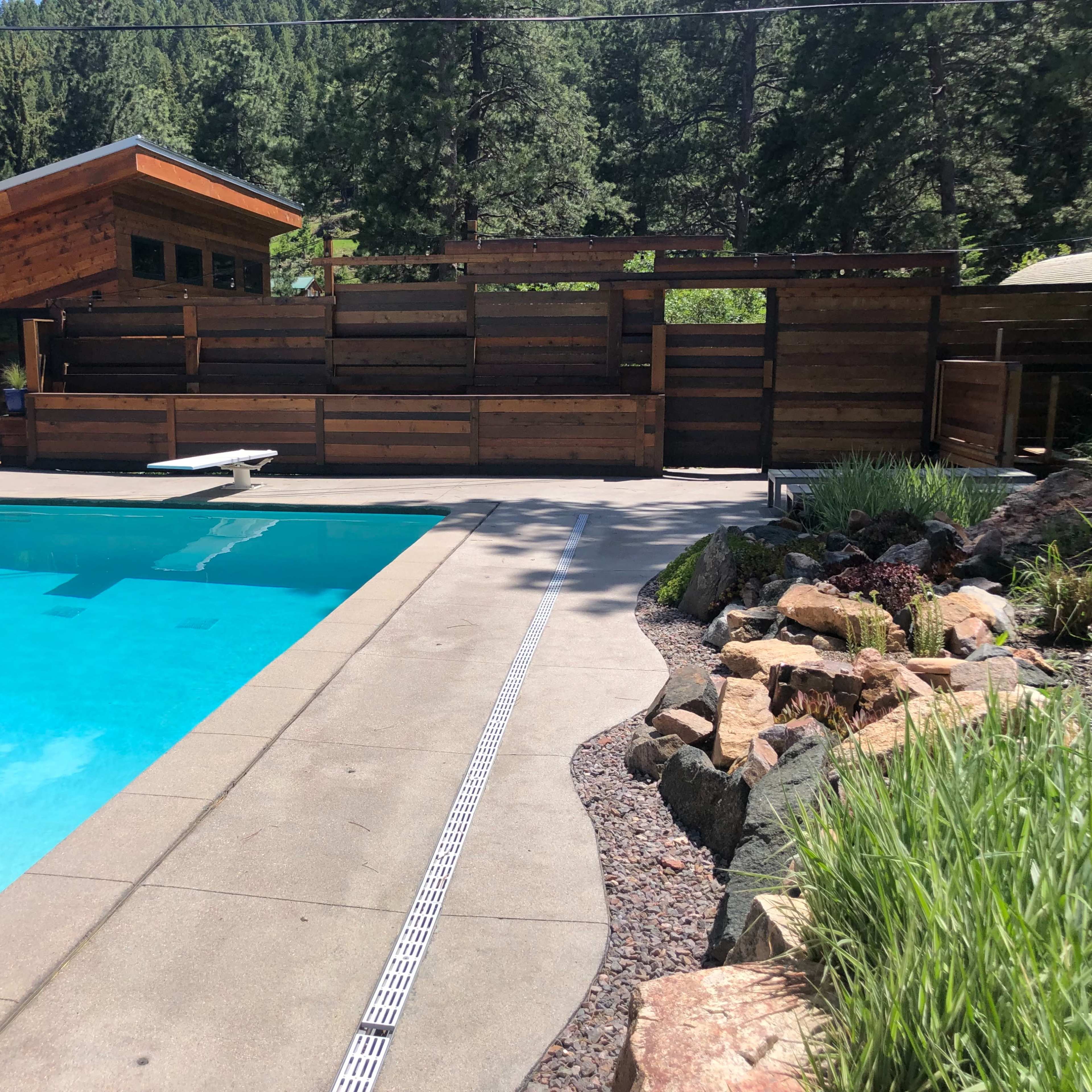 A rectangular swimming pool beside a wooden deck and modern cabin set against a backdrop of trees.