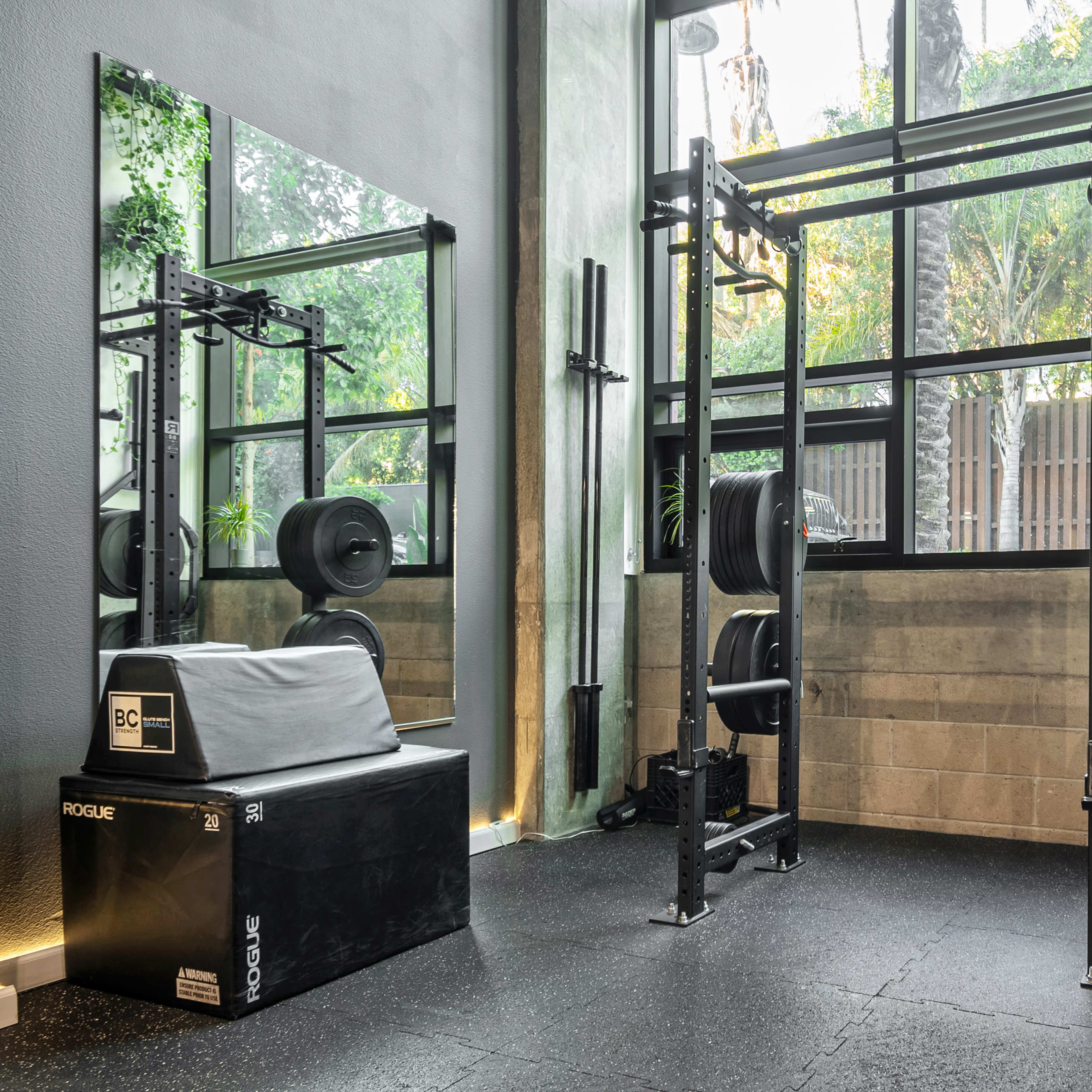 A modern gym area featuring a weightlifting rack, a black exercise box, and various weight plates in a bright space with large windows.