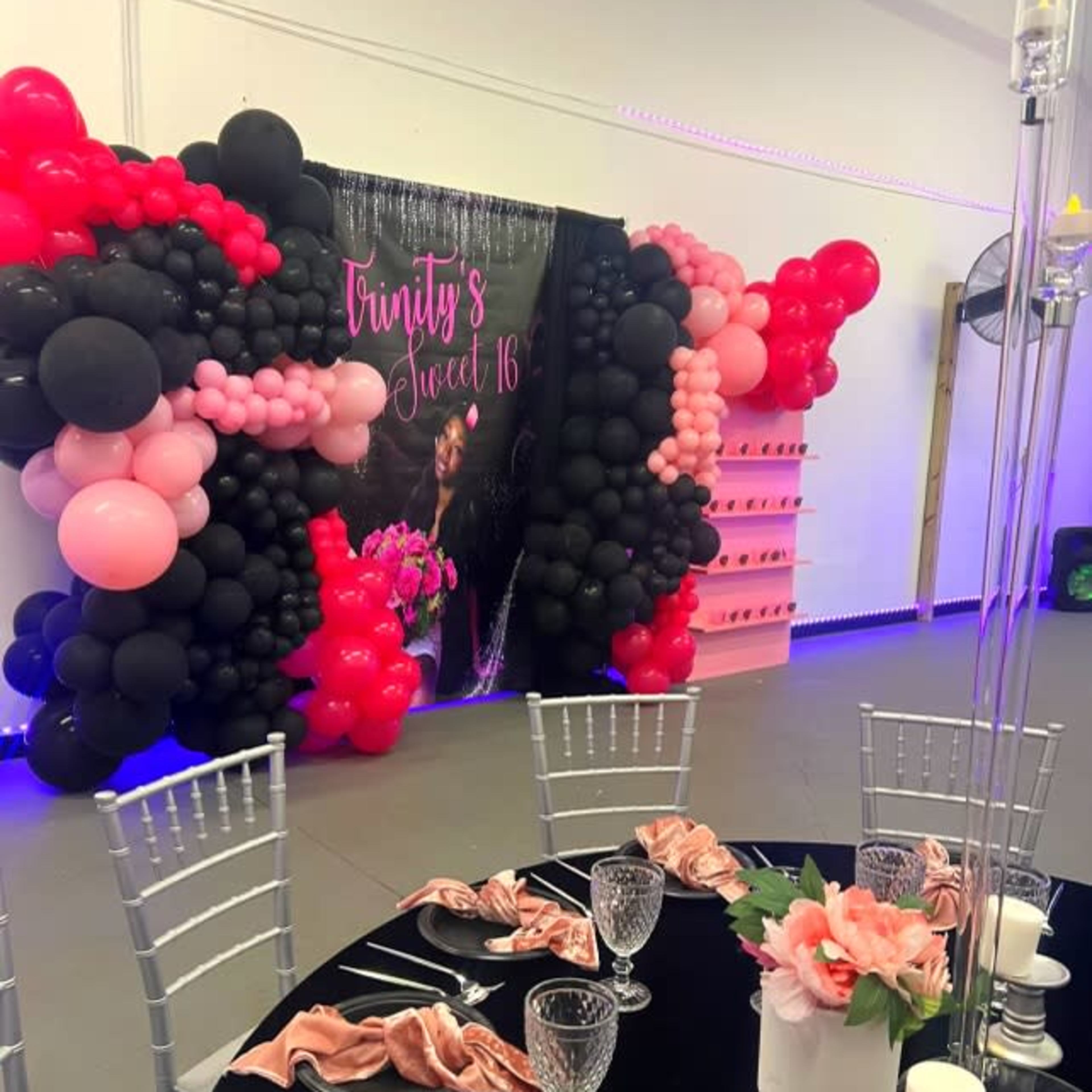 A decorated event space for a Sweet 16 celebration, featuring a wall of black, pink, and red balloons, a large banner, and a table set with plates, glasses, and napkins.