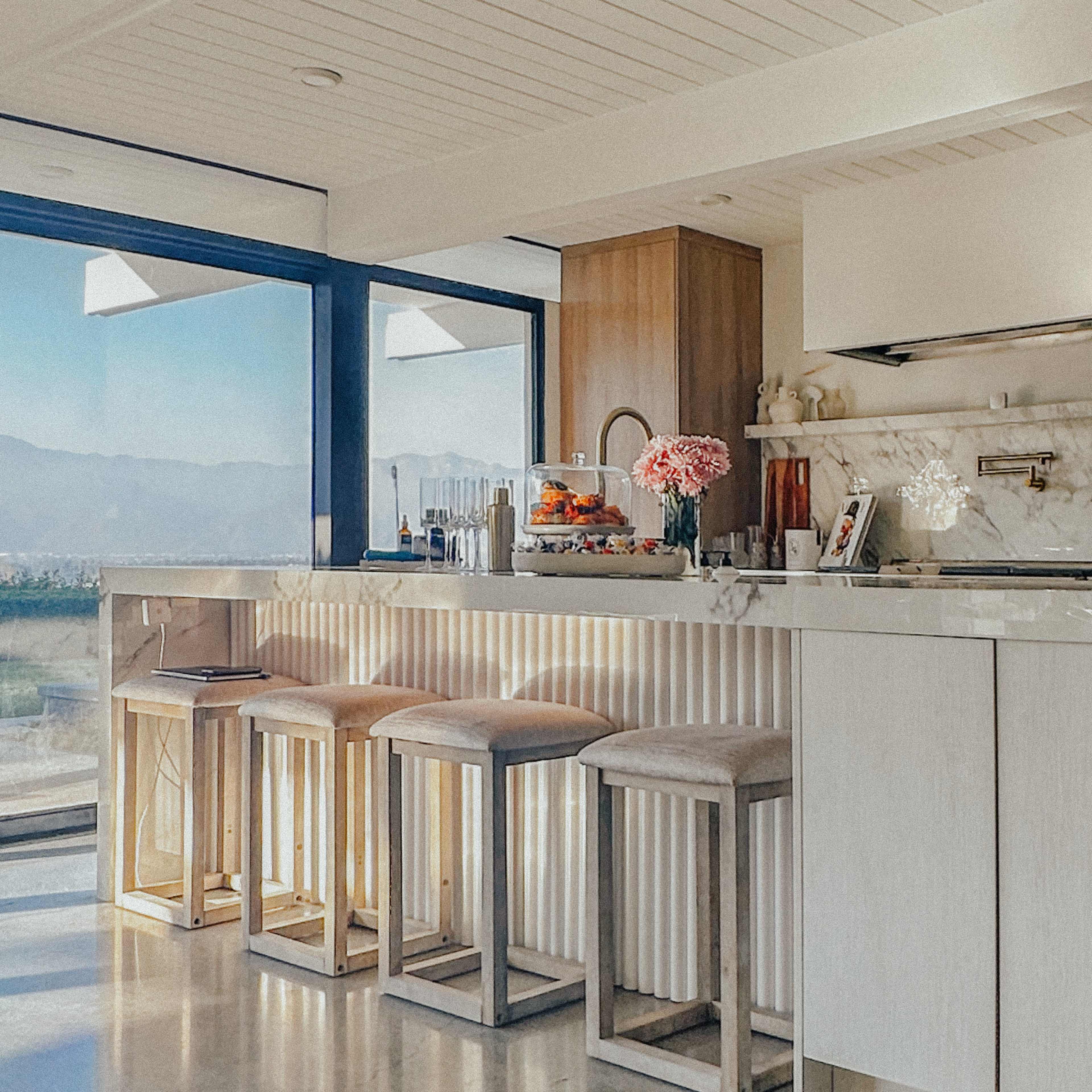 A modern kitchen features a marble countertop with bar stools, large windows allowing natural light, and a view of the outdoors.