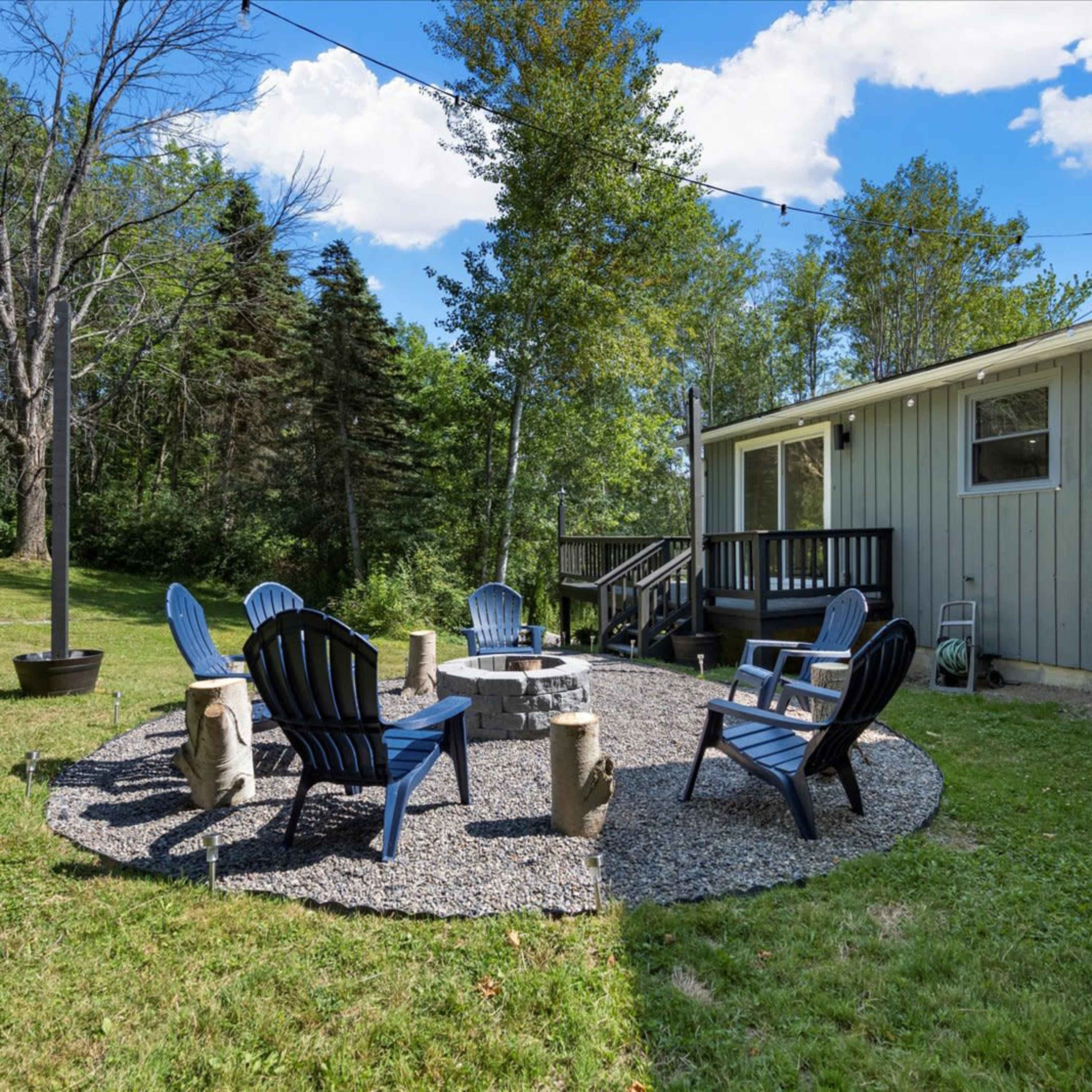 A circular seating area with blue chairs surrounds a stone fire pit in a grassy outdoor space next to a house.