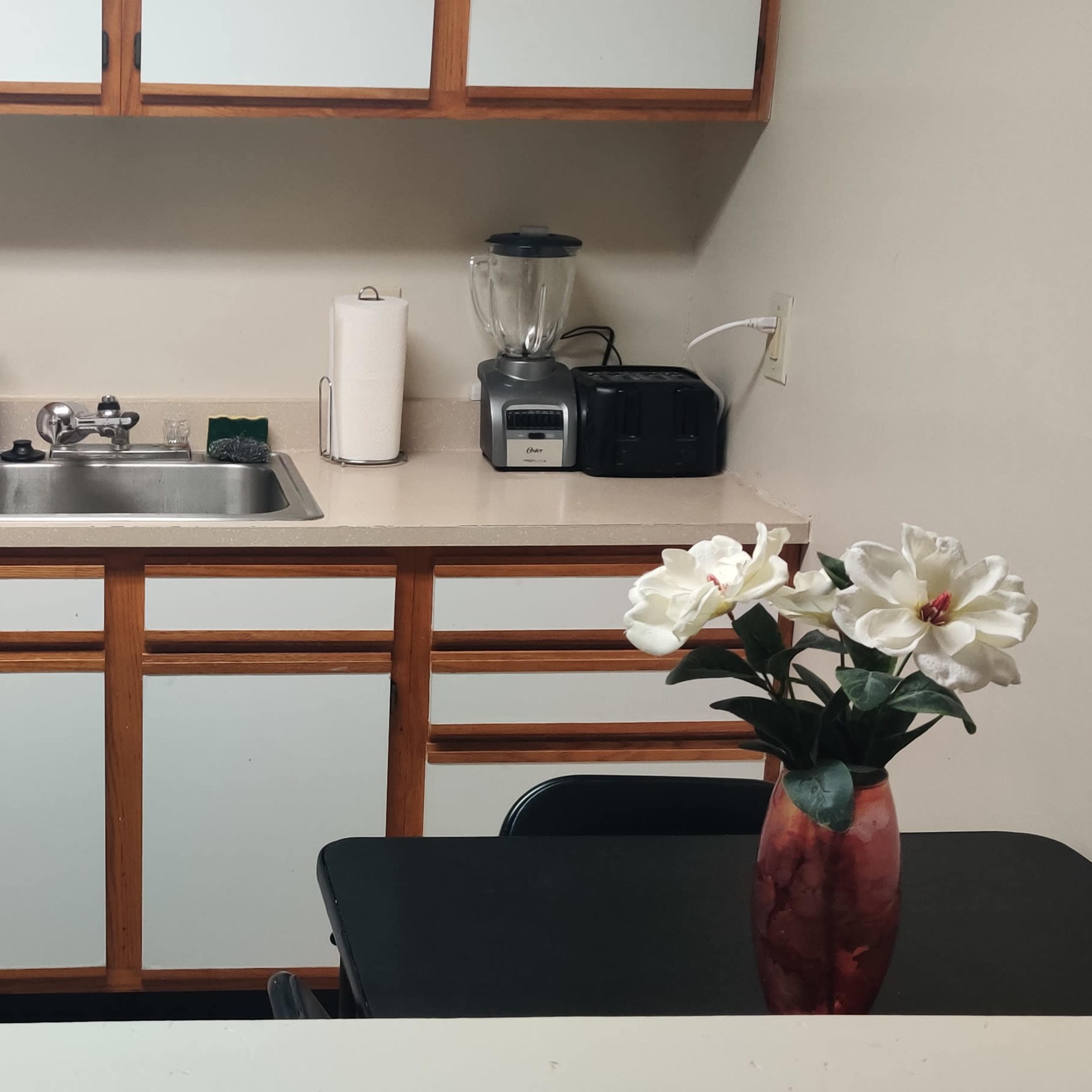 A small kitchen features a white refrigerator, stove, microwave, and sink with a blender, alongside a table with a vase of flowers.