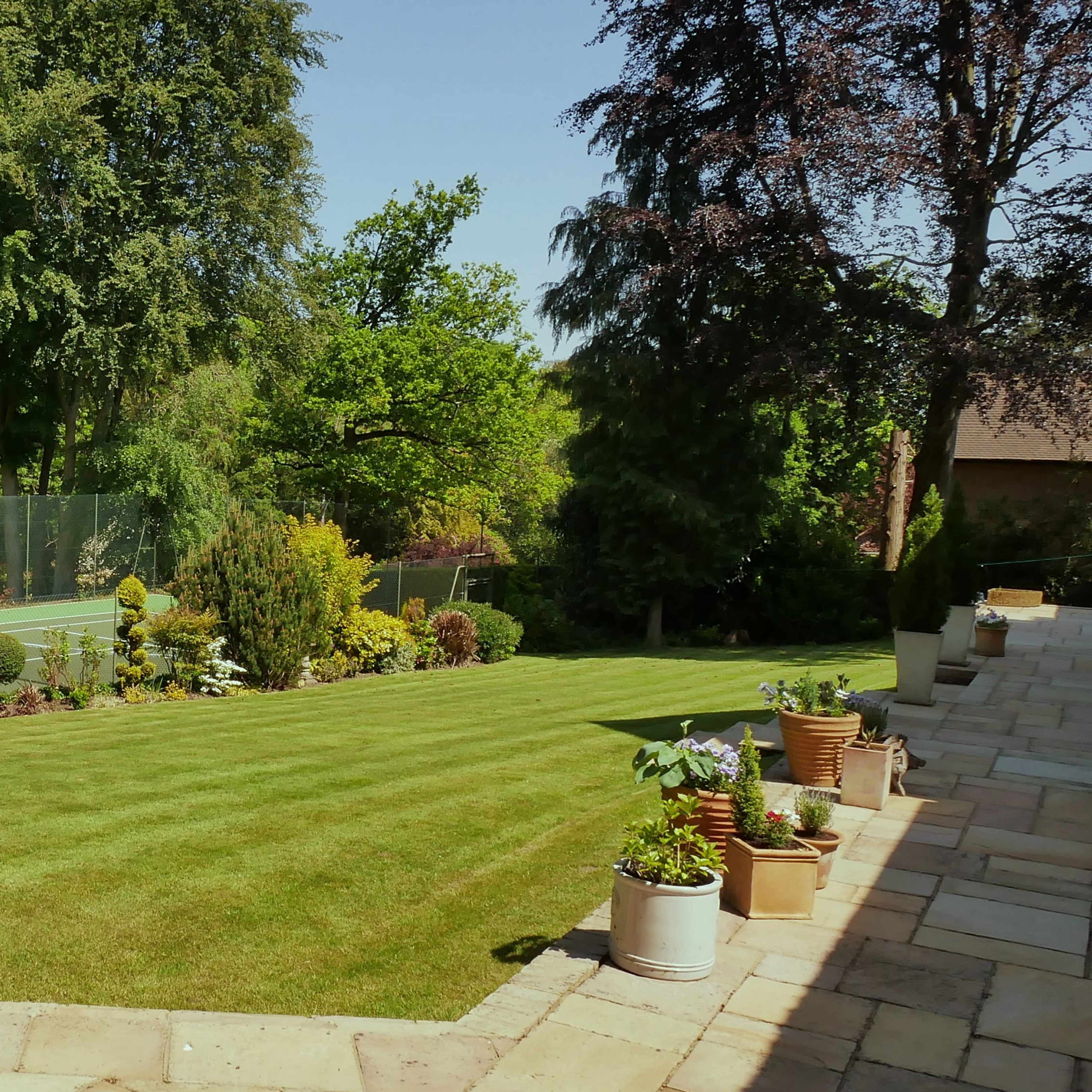 A well-maintained garden with a neatly trimmed lawn, decorative plants, and a tennis court in the background.