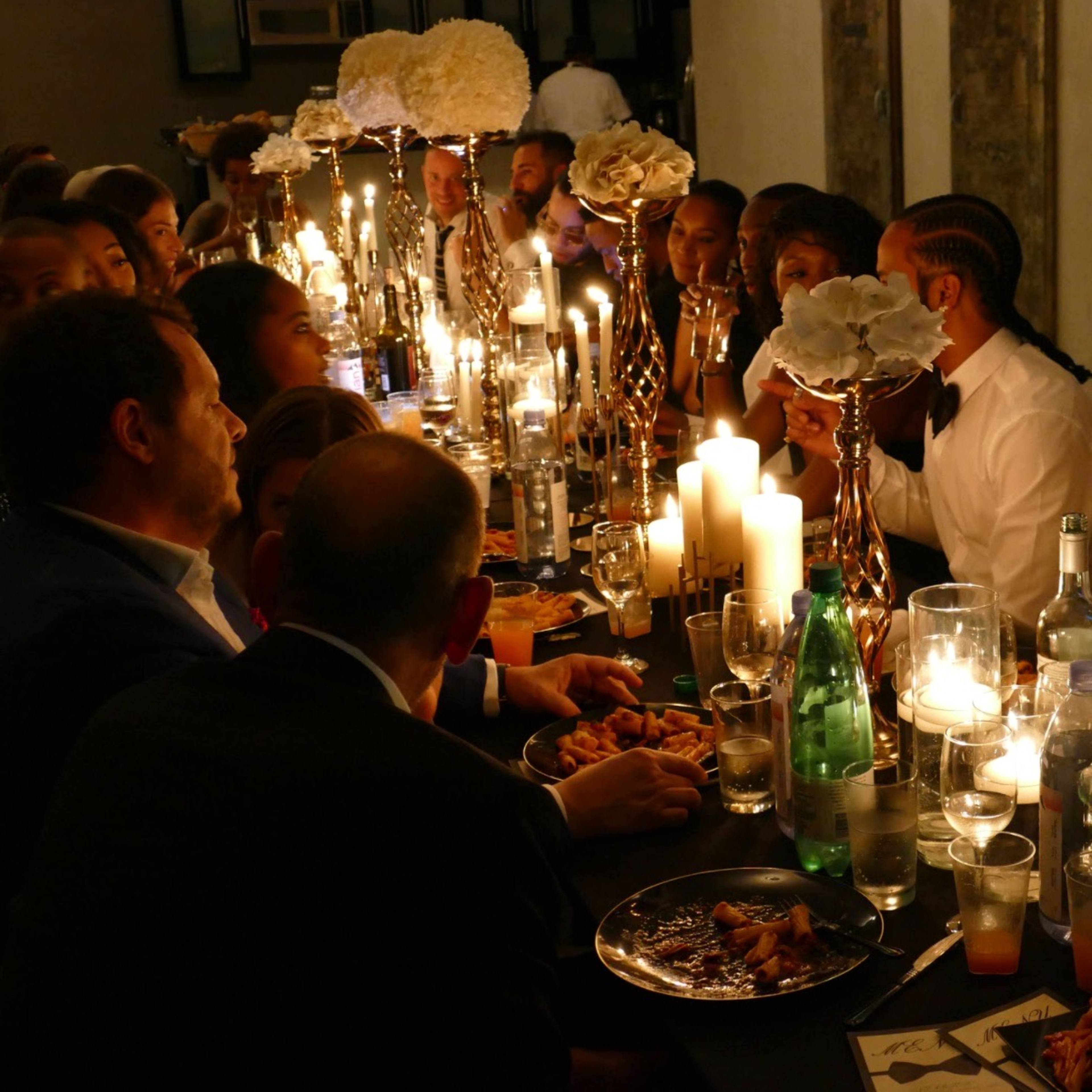 A large, elegantly set dining table is filled with guests enjoying a formal dinner, illuminated by multiple candles and floral centerpieces.