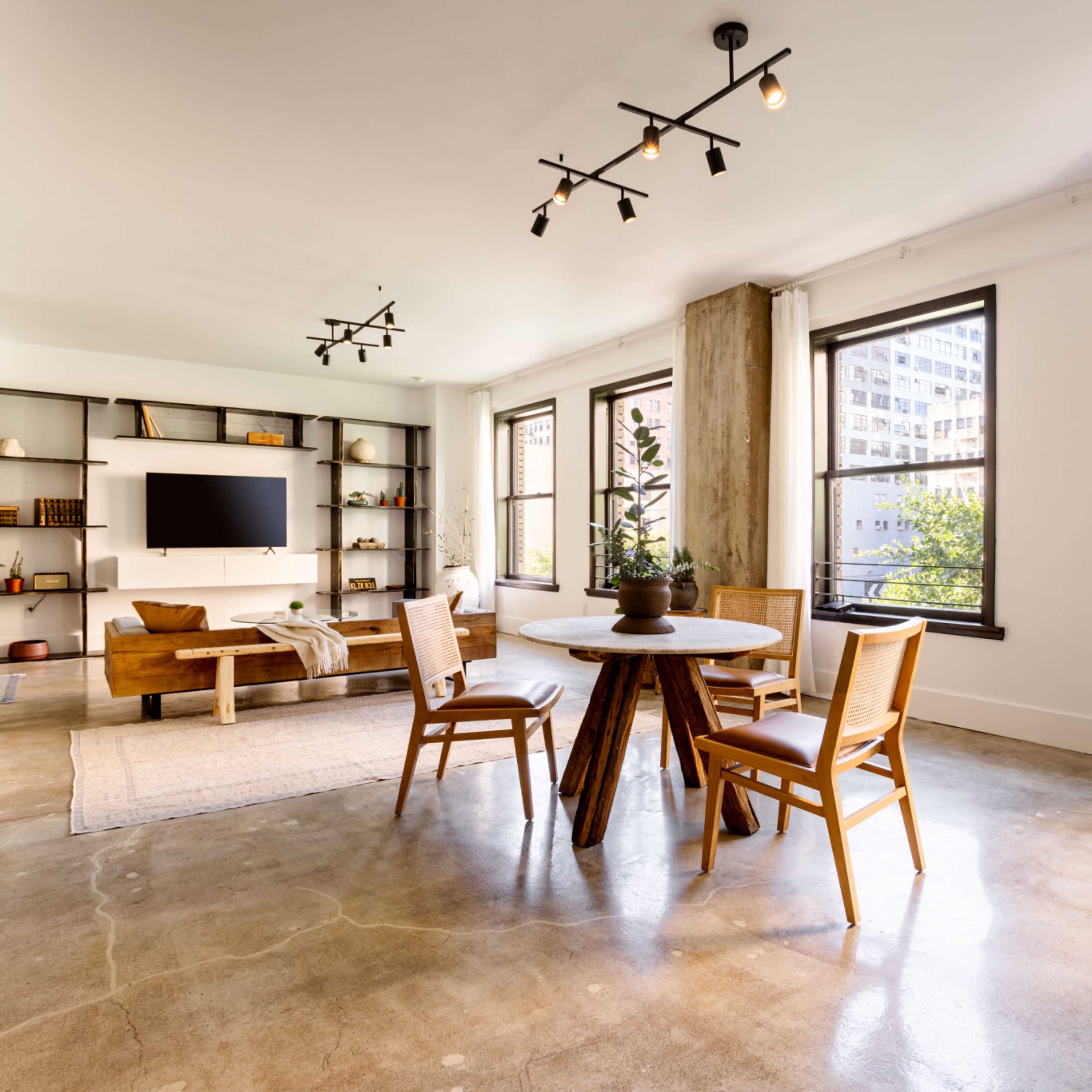 A modern living space featuring a circular dining table surrounded by chairs, a sofa, and a TV shelf against the wall, with large windows allowing natural light to enter.