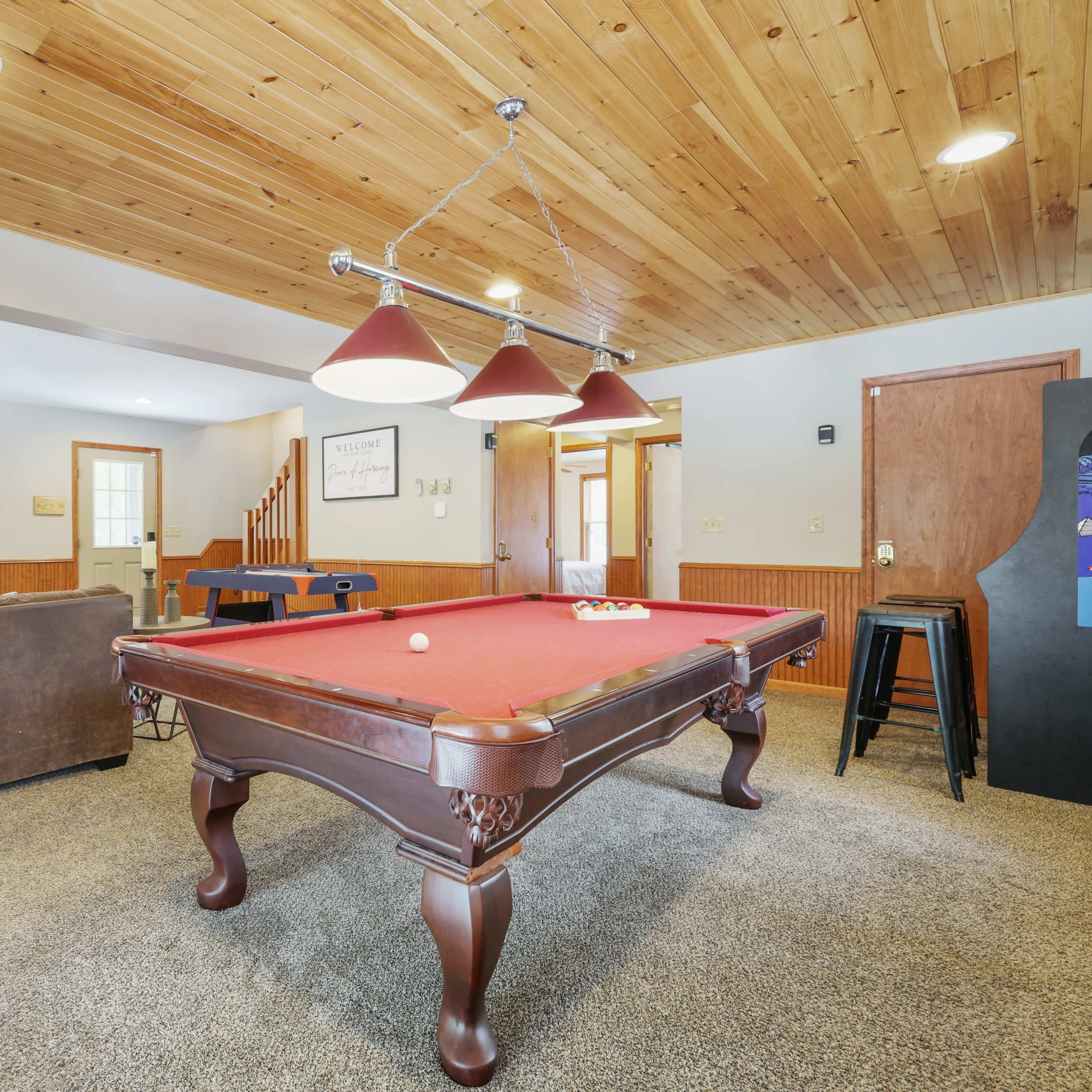 A billiard table with red felt is situated in a room featuring a wooden ceiling, a brown couch, and a television.