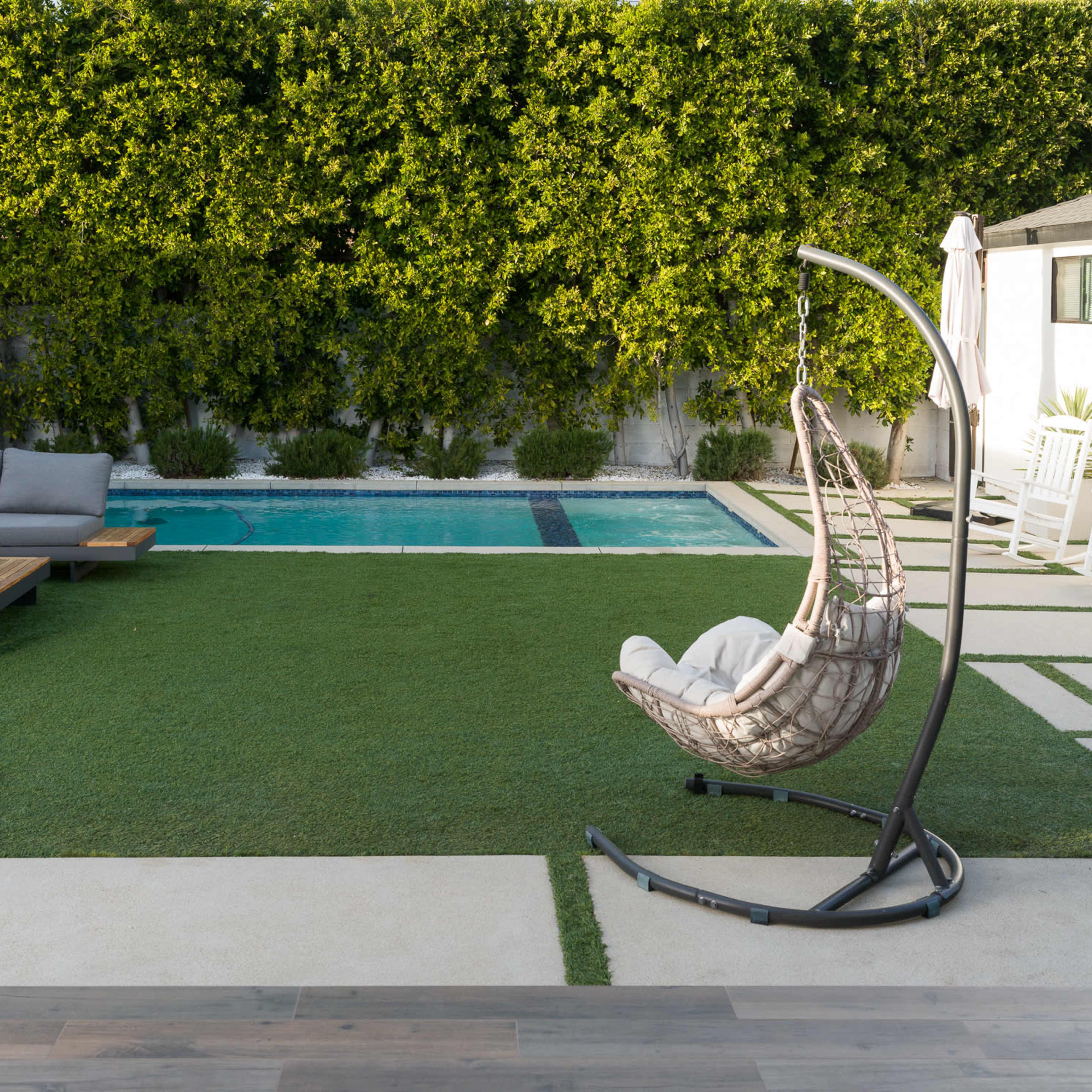 A modern backyard featuring a swimming pool, a large sectional sofa, and a hanging chair on a grassy area.