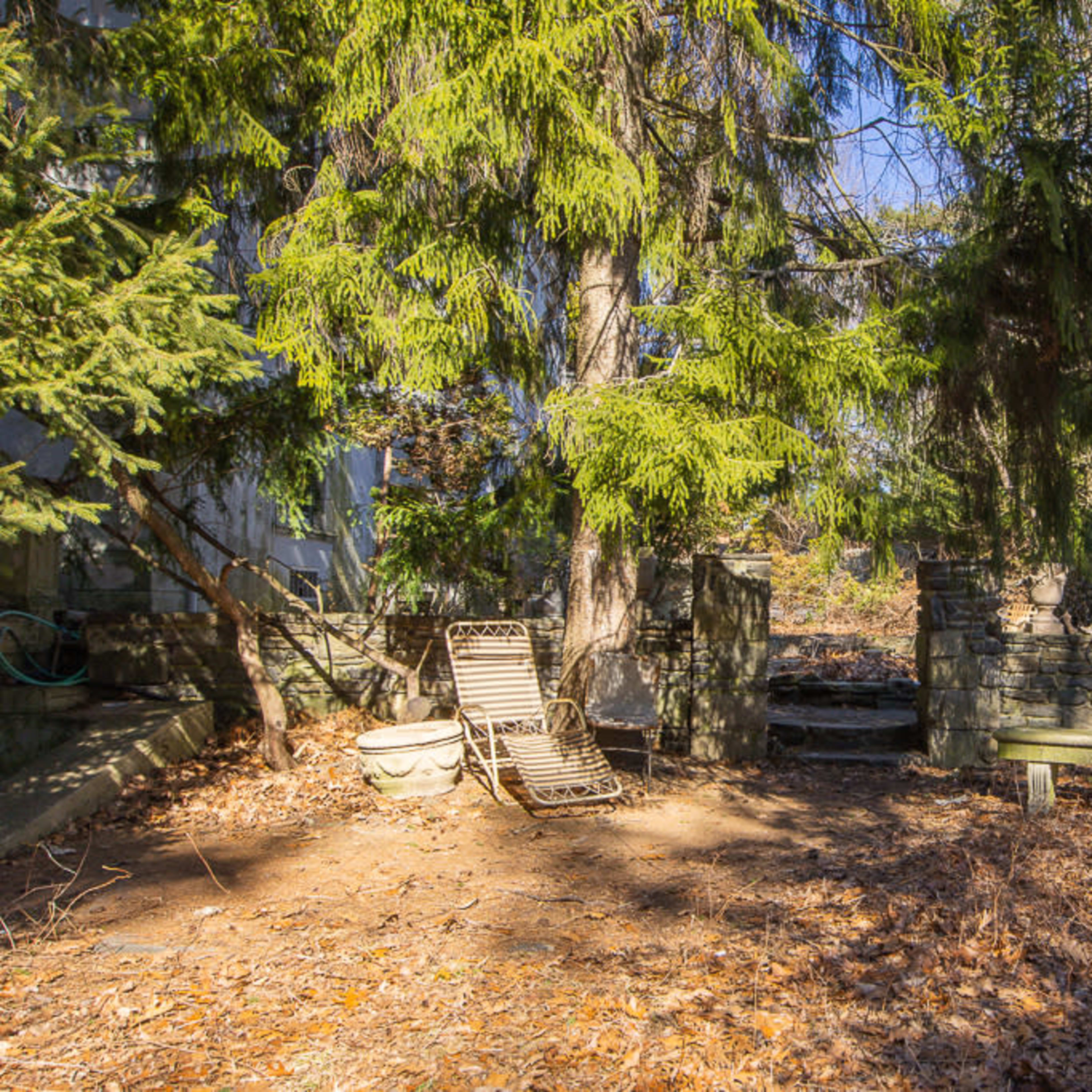 A serene outdoor space with two wooden chairs positioned on a leaf-covered ground, surrounded by tall trees and stone walls.