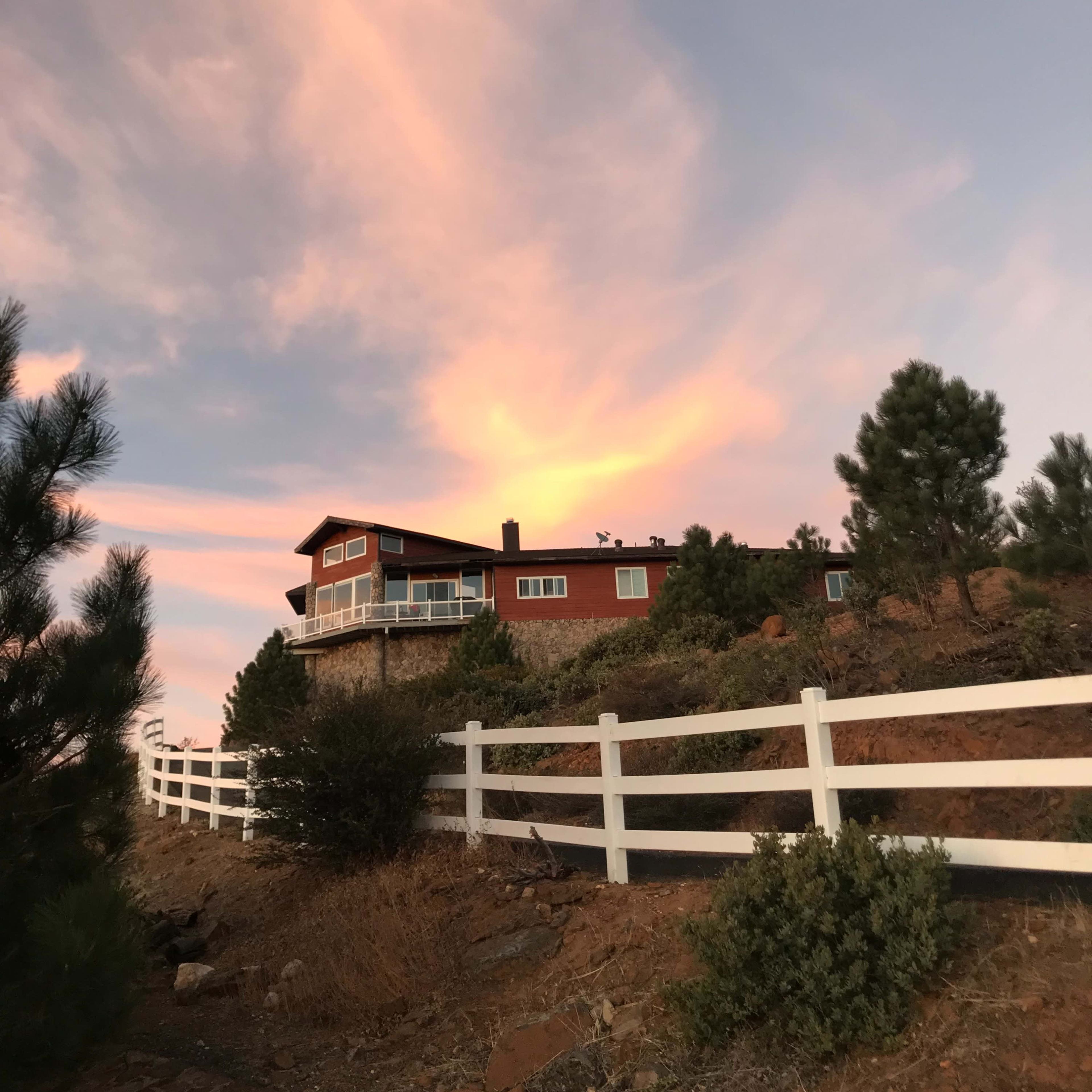 A house with a deck is situated on a hillside against a colorful sky at sunset, surrounded by trees and a white fence.
