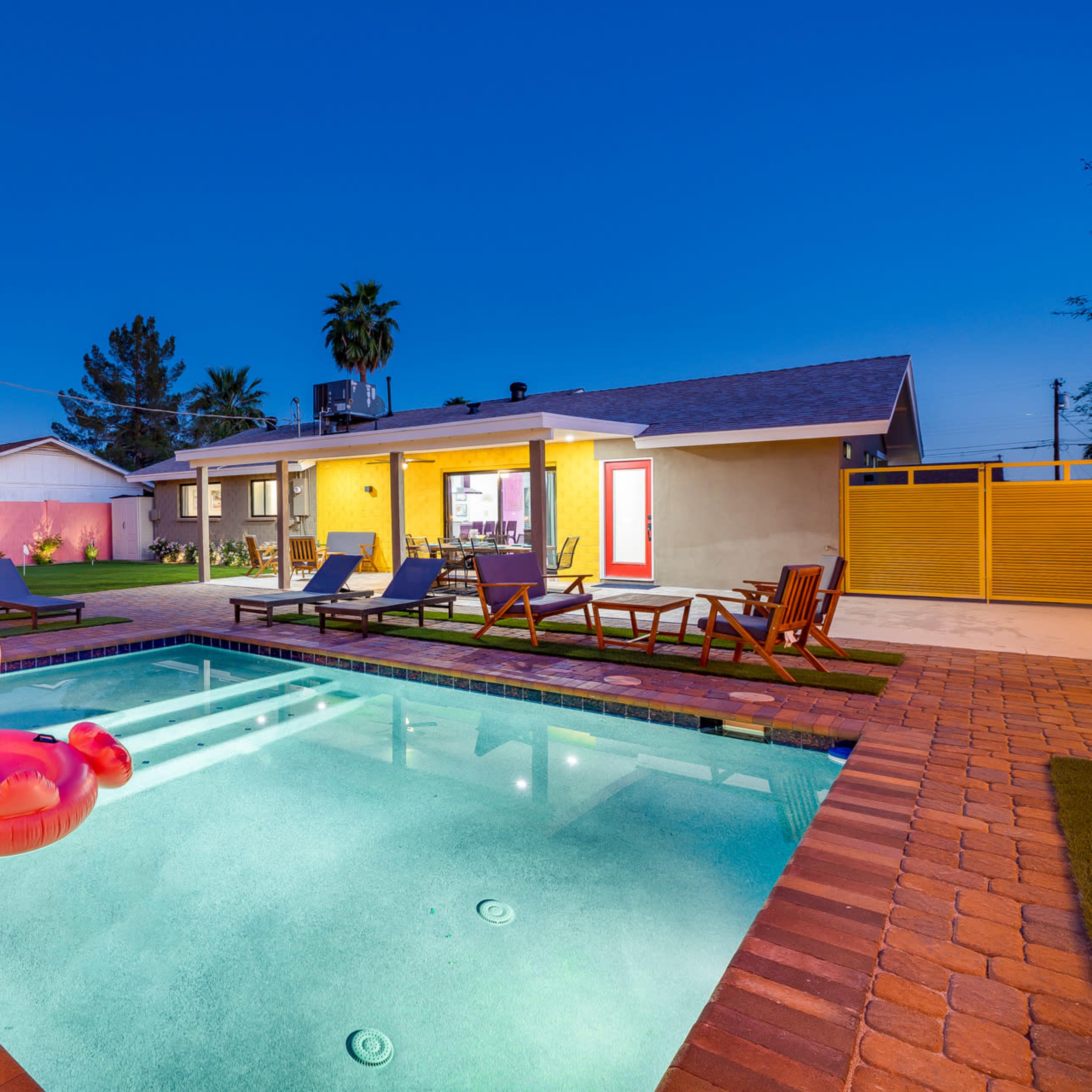 A swimming pool with a pink flamingo float is surrounded by lounge chairs and a well-lit backyard at dusk.