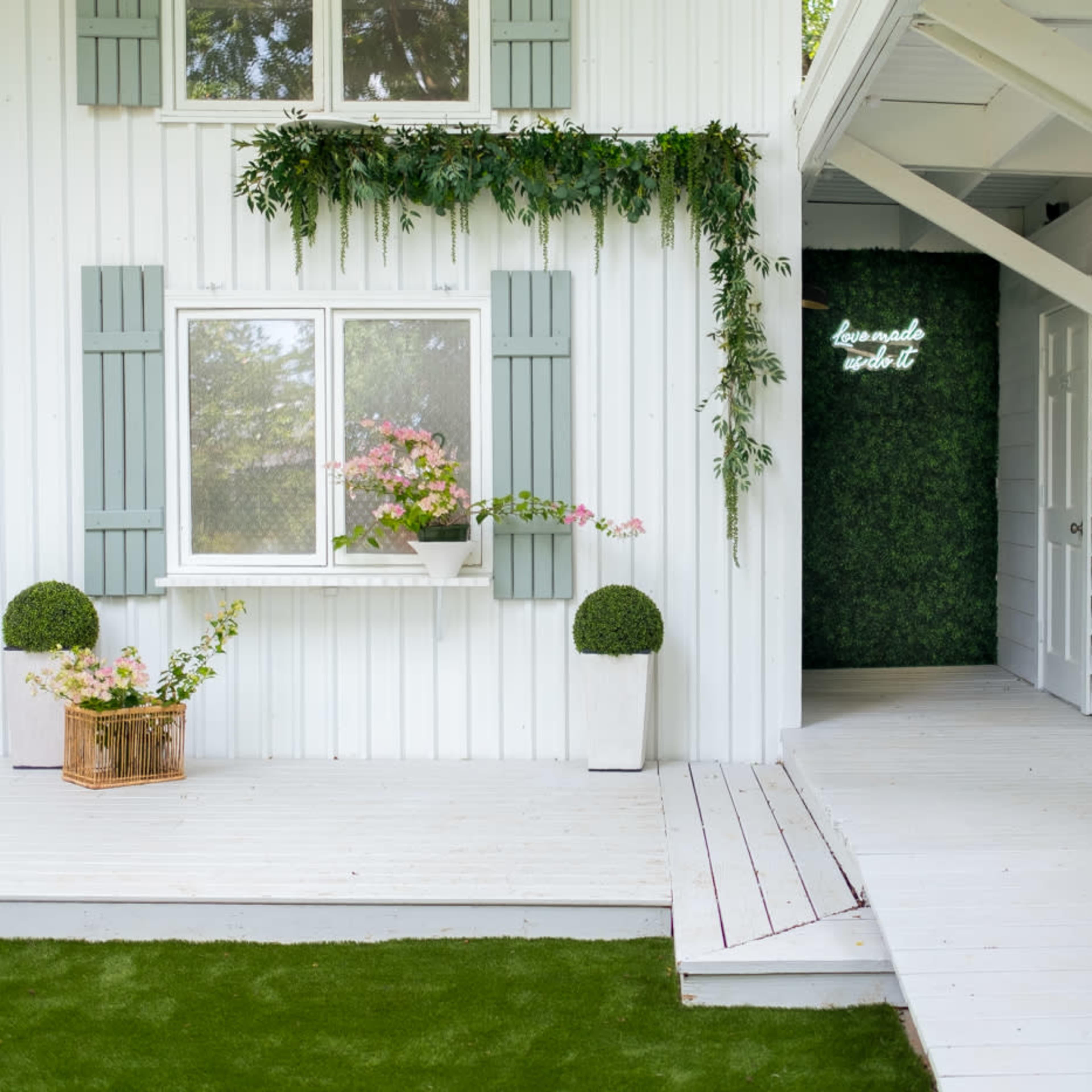A white wooden house with green shutters, a flower-filled window box, and a porch leading to a door framed by a wall of greenery.