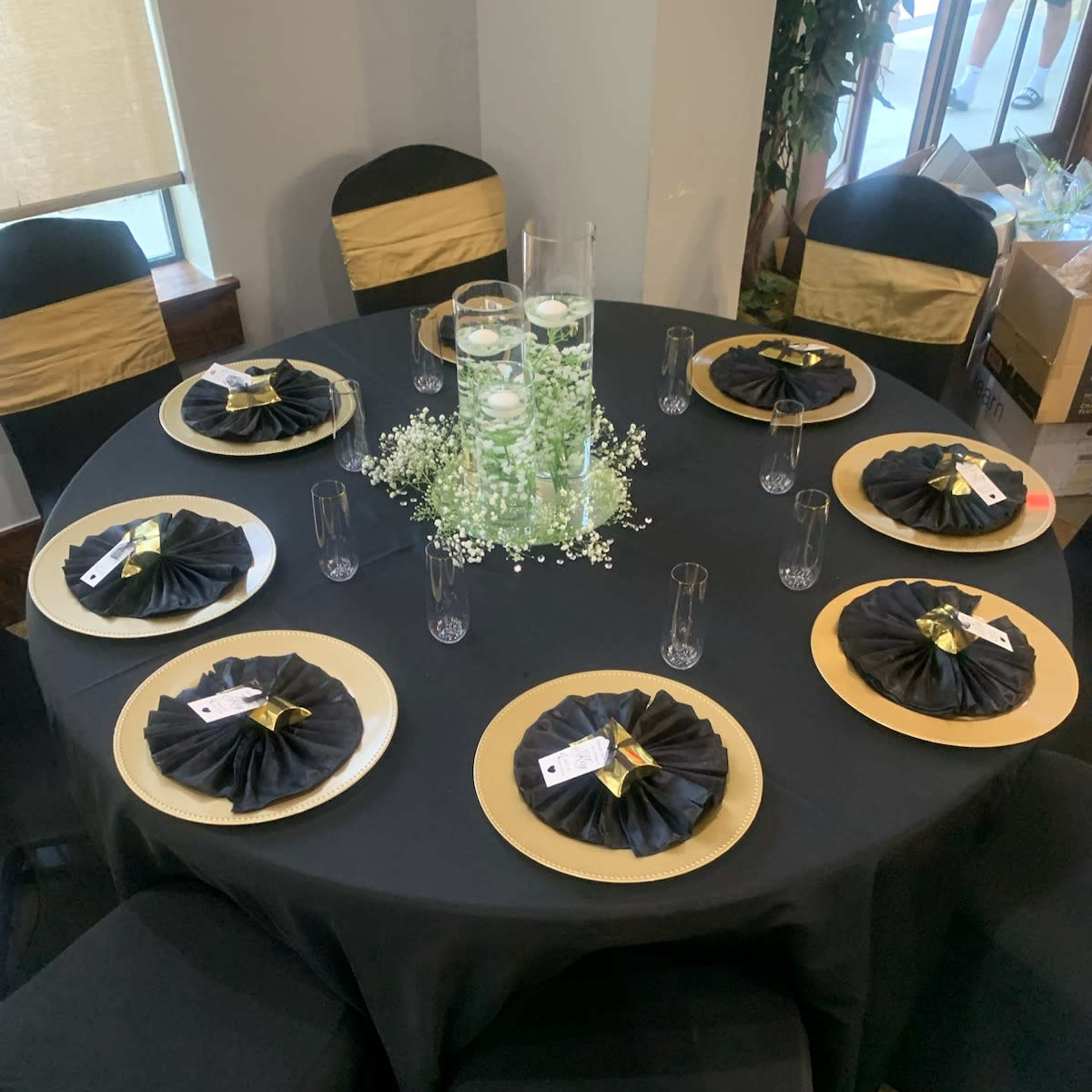 A round table is set with black and gold decorations, featuring plates with black napkins and a centerpiece of candles surrounded by greenery.