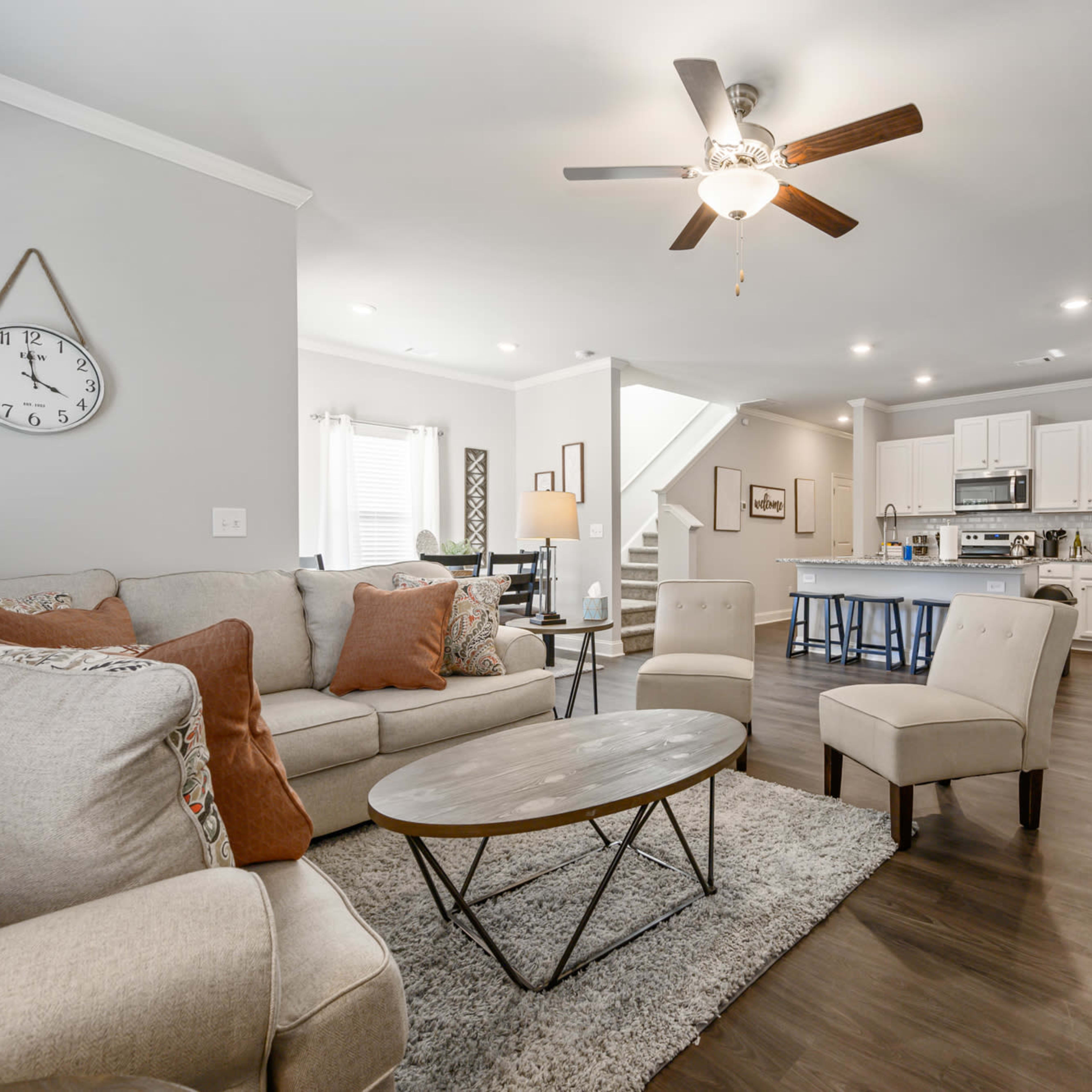 A modern living room with a light-colored sofa, a round coffee table, and a ceiling fan, leading into a kitchen with white cabinets and a dining area.