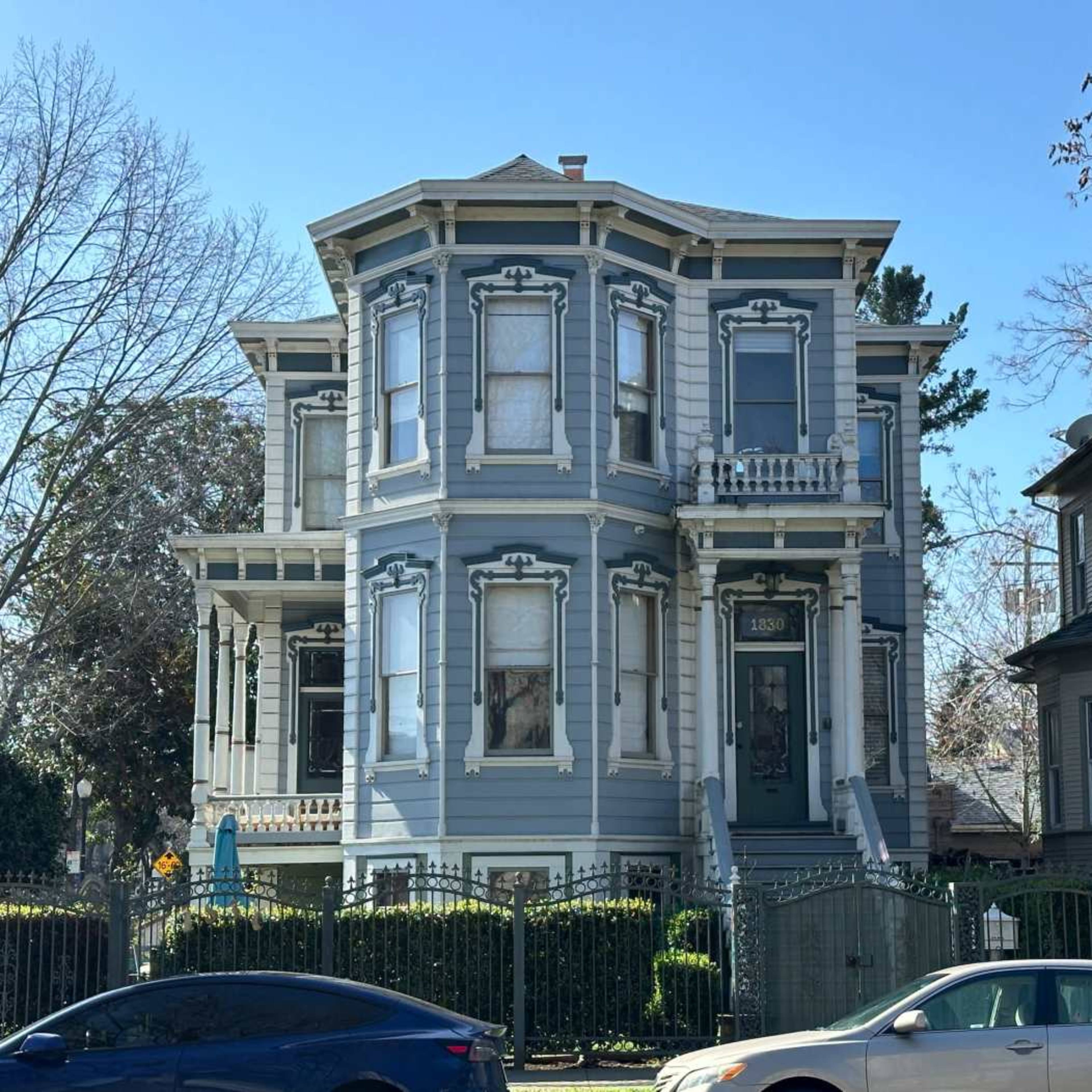 A large, two-story Victorian-style house painted in light blue, featuring intricate architectural details and a well-maintained garden.
