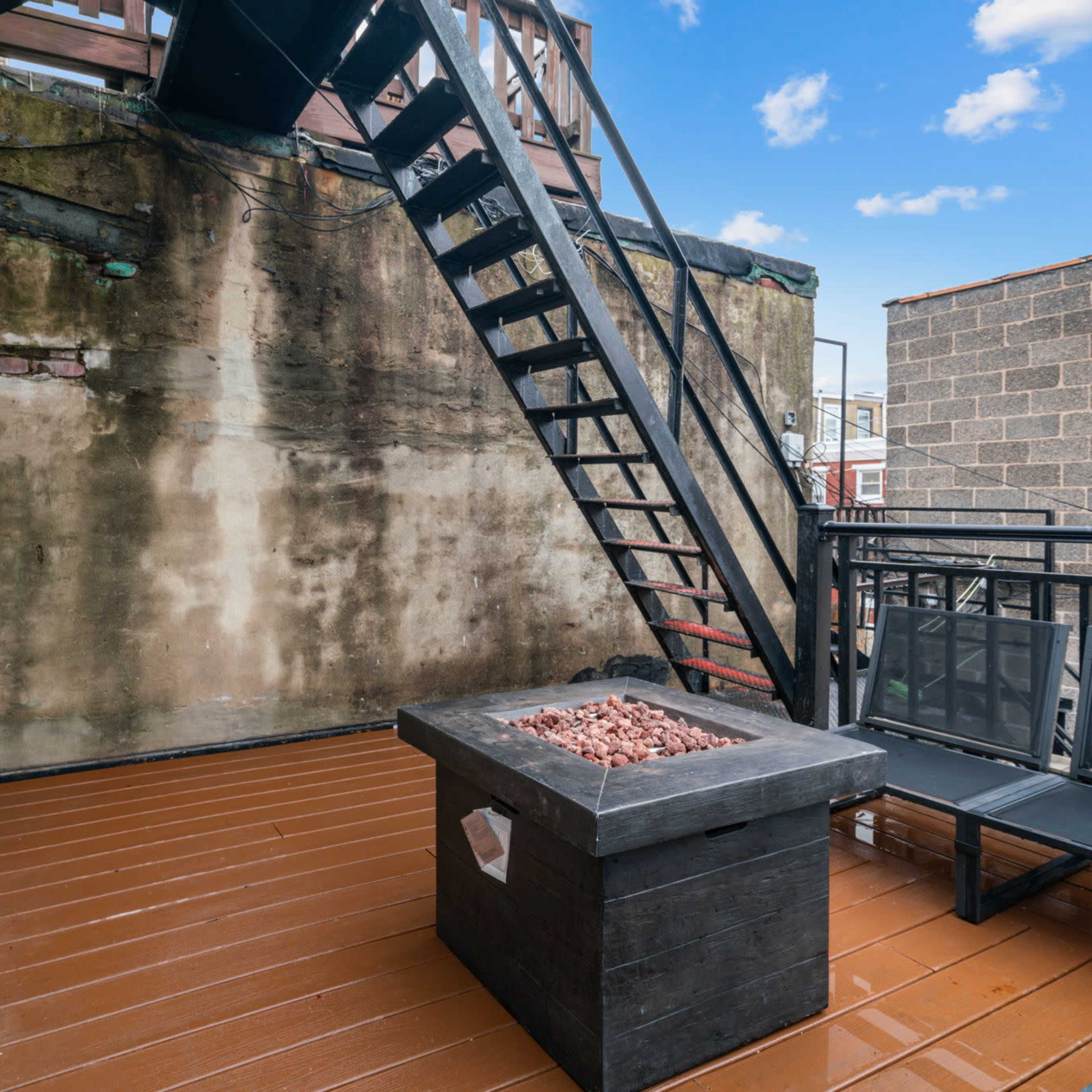 A rooftop balcony with a wooden deck, an outdoor seating area, and a metal staircase leading to an upper level.