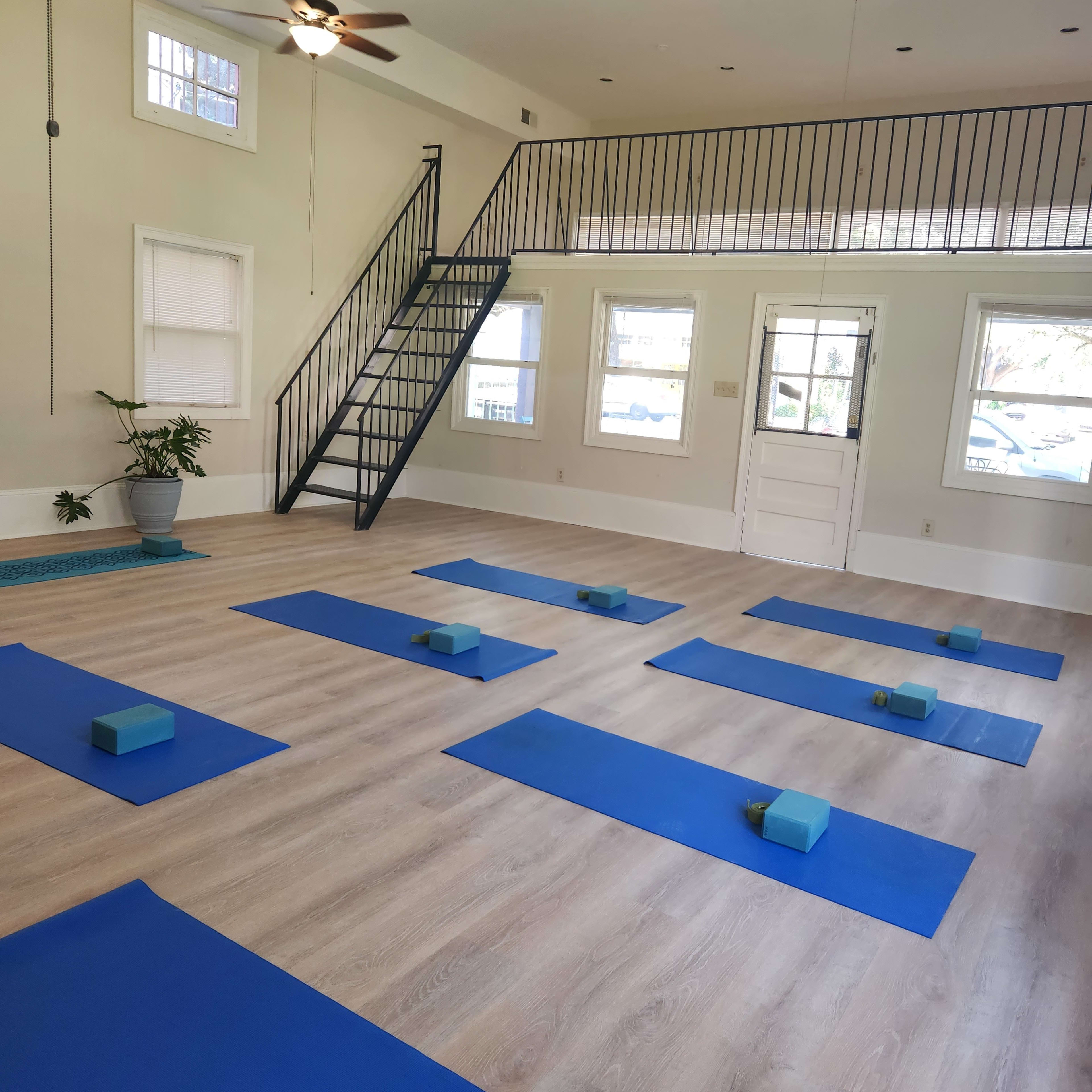 A spacious yoga studio with multiple blue mats and blocks arranged on the wooden floor, illuminated by ceiling fans and natural light from the windows.