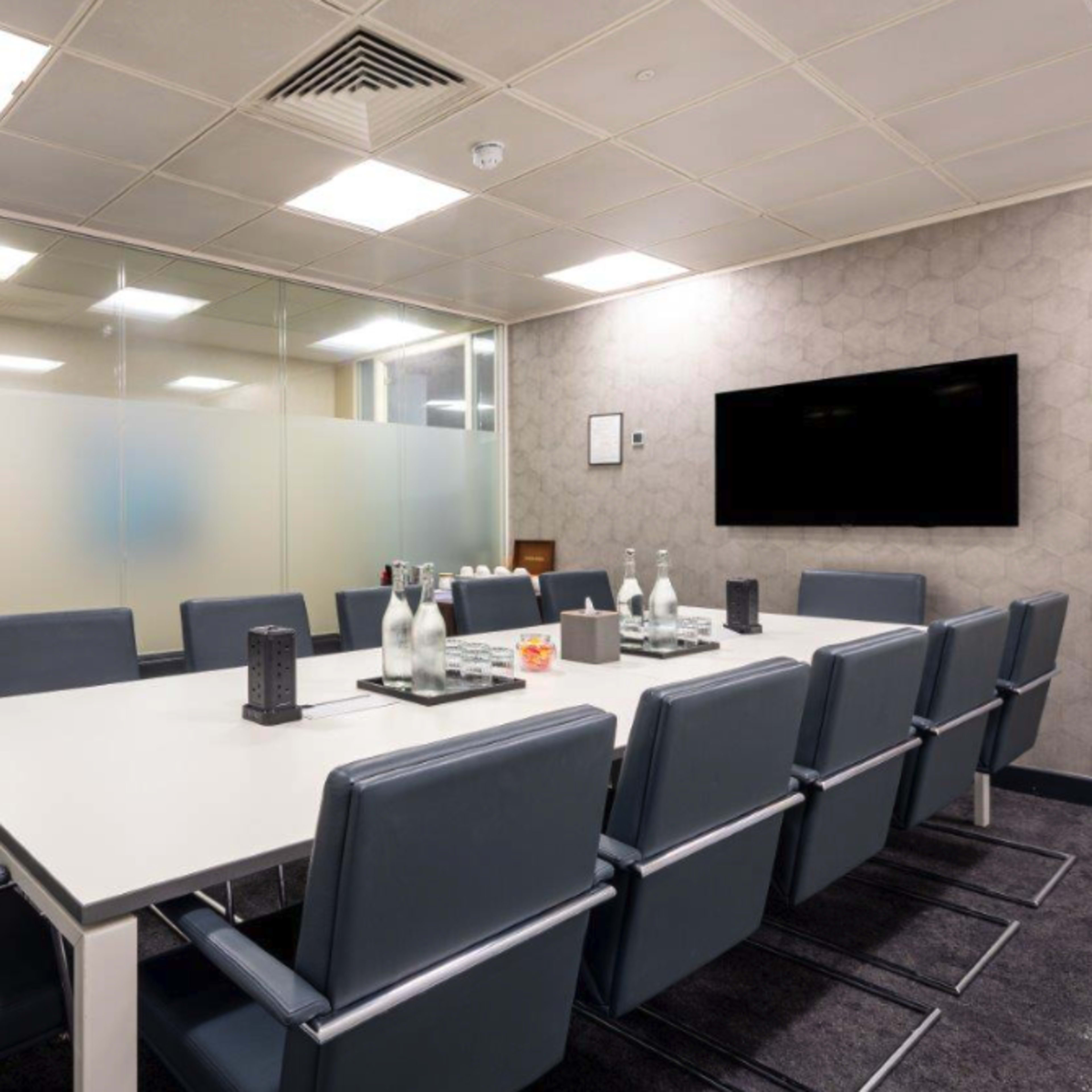 A modern conference room with a long table surrounded by several leather chairs, glass walls, and a wall-mounted television.