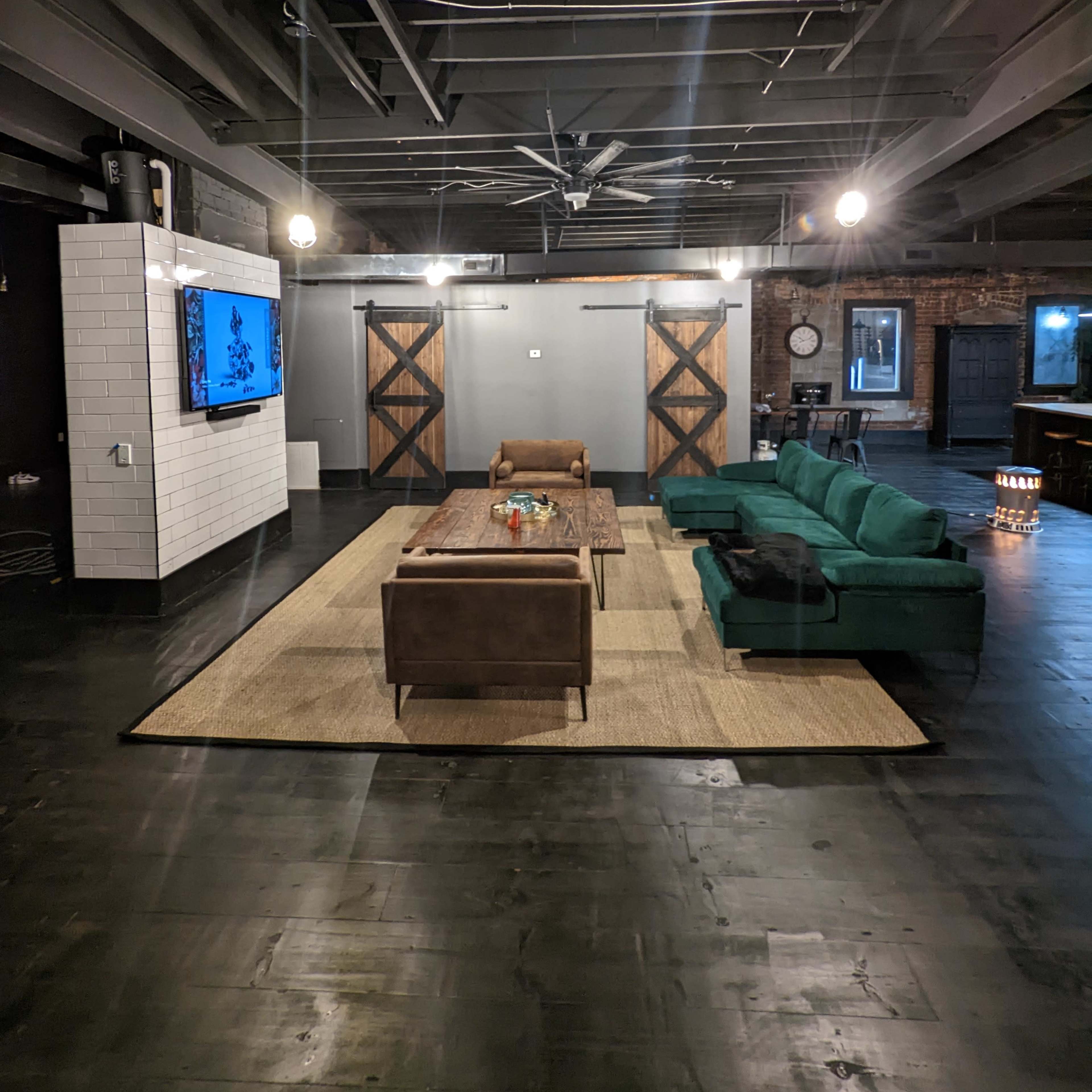 A spacious living area features a green sectional sofa and a wooden coffee table, with a large television mounted on a white wall and exposed brick accents.