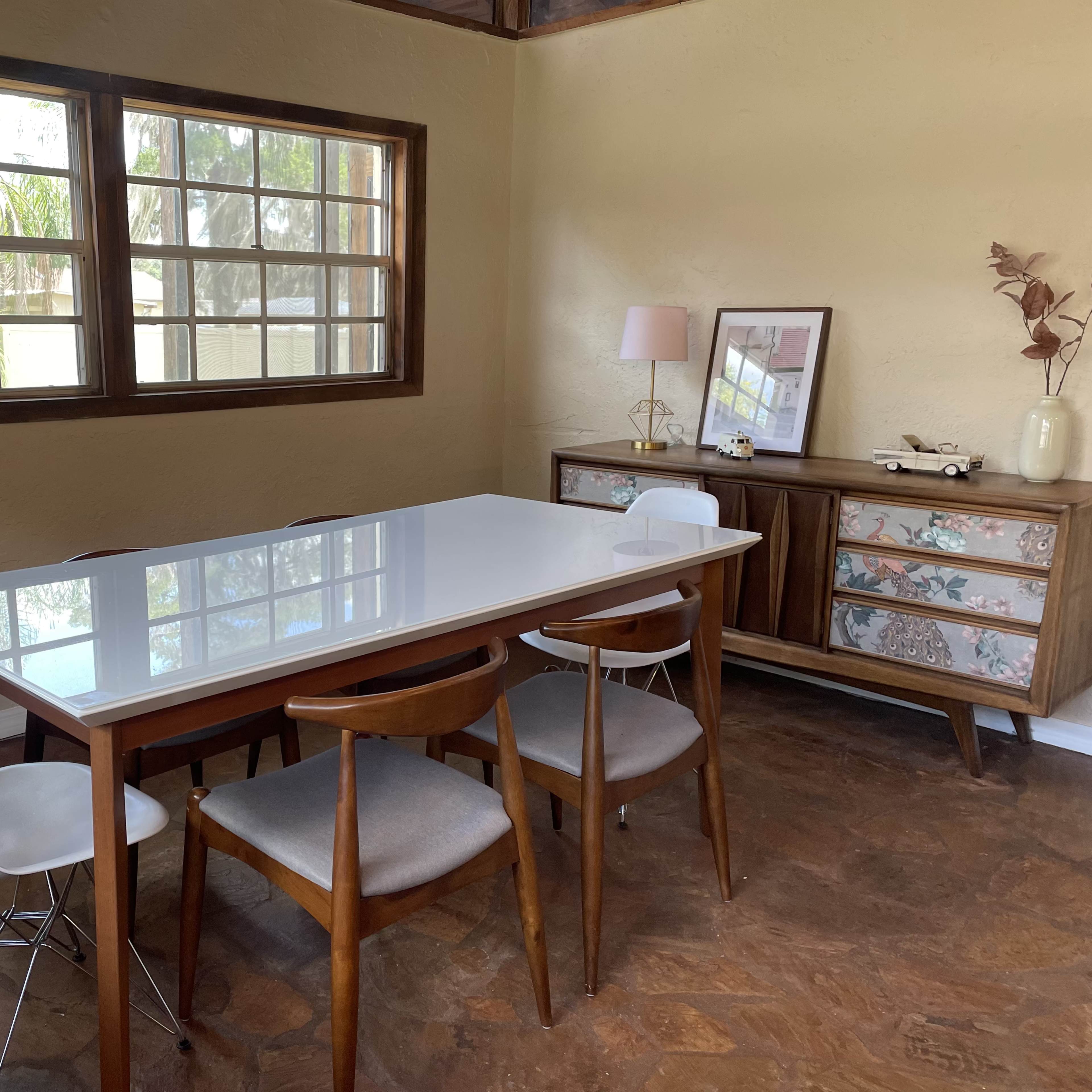 A dining area features a white table surrounded by four wooden chairs, with a cabinet and a window displaying greenery outside.
