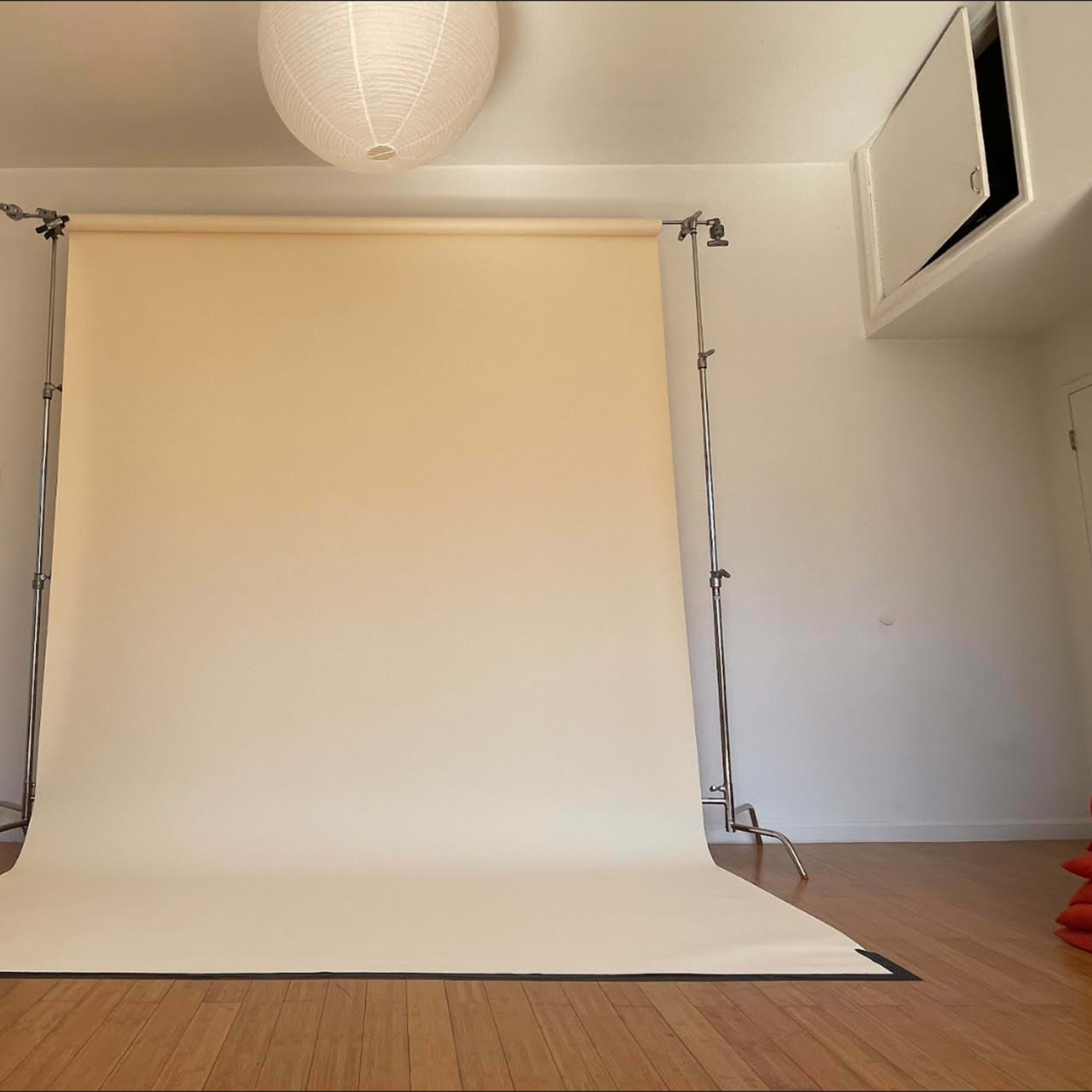A minimalist photography studio with a beige backdrop set on a wooden floor, illuminated by a pendant light.