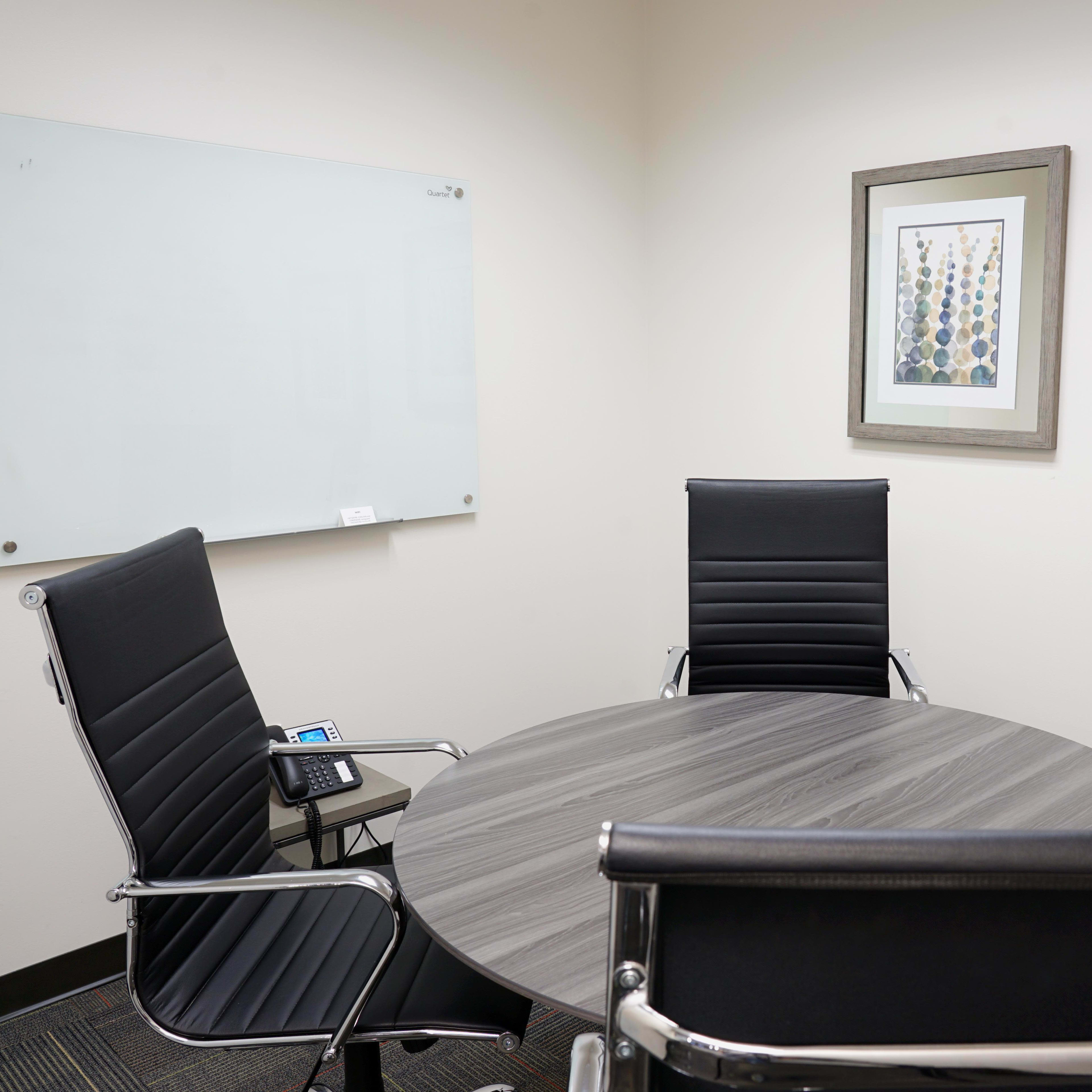 A small conference room features a circular table surrounded by four black chairs, with a whiteboard on one wall and two framed artworks nearby.