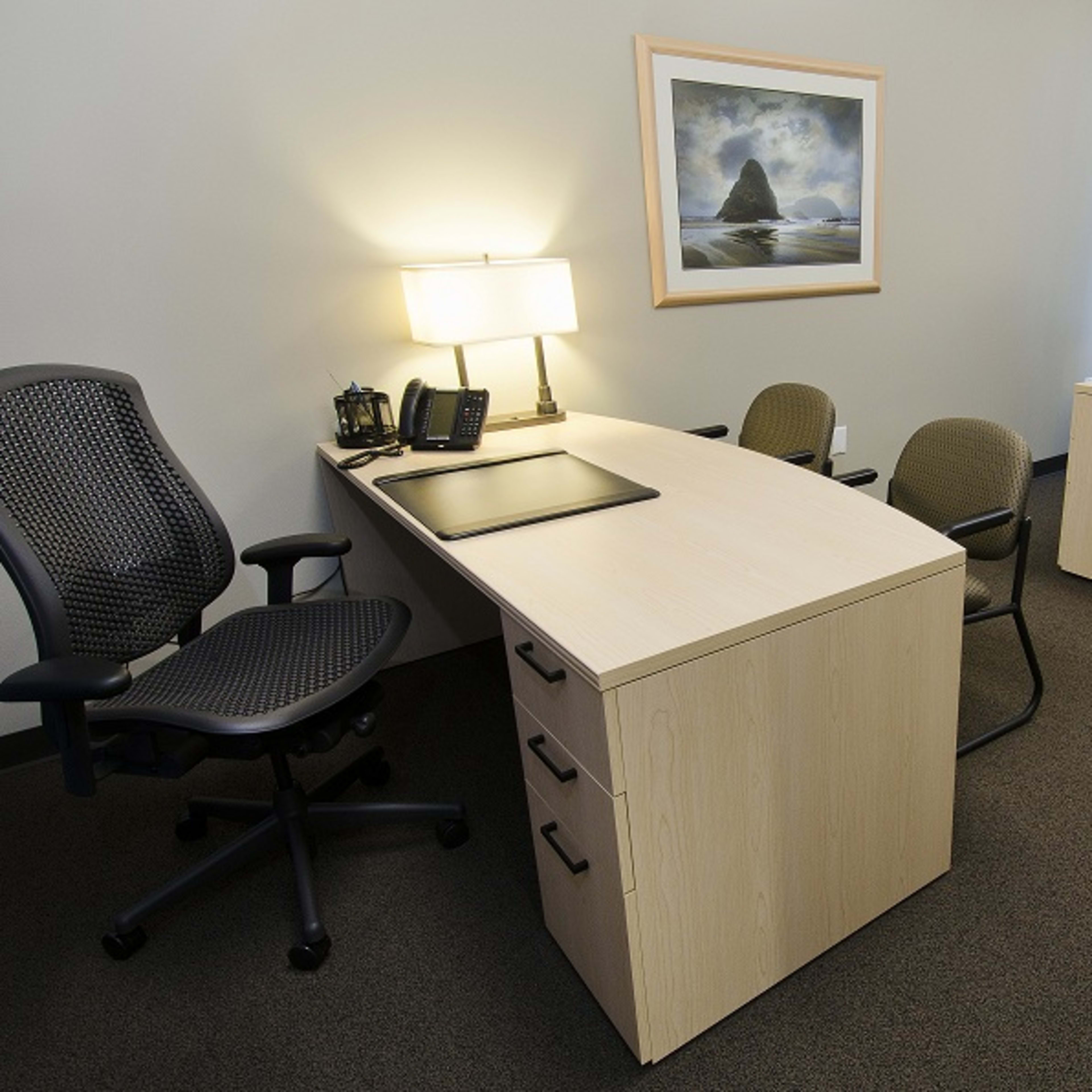 A modern office space with two desks, an office chair, and a waiting area with stackable chairs, all illuminated by lamps and natural light.