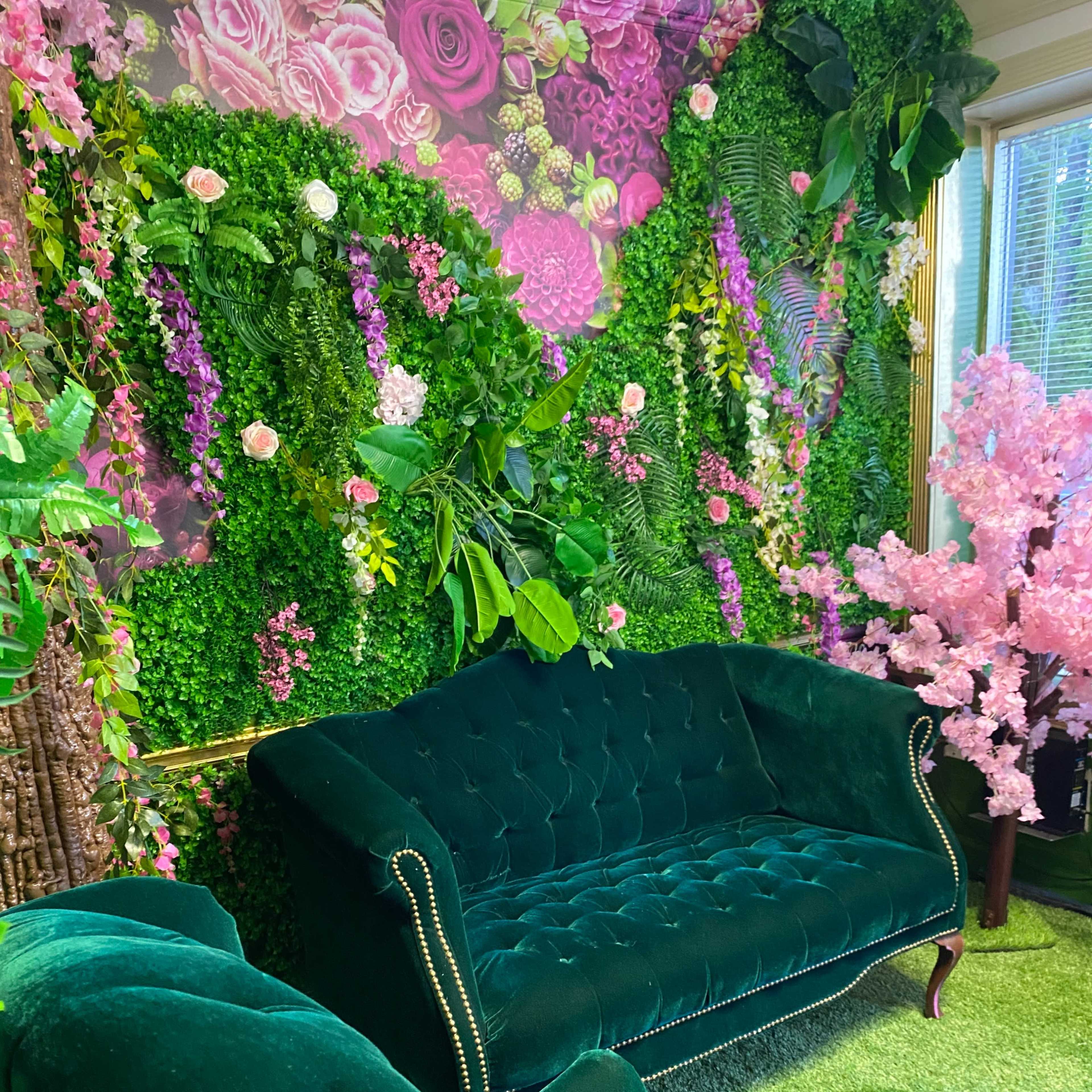 A green velvet couch and armchair arranged in front of a floral and greenery wall.