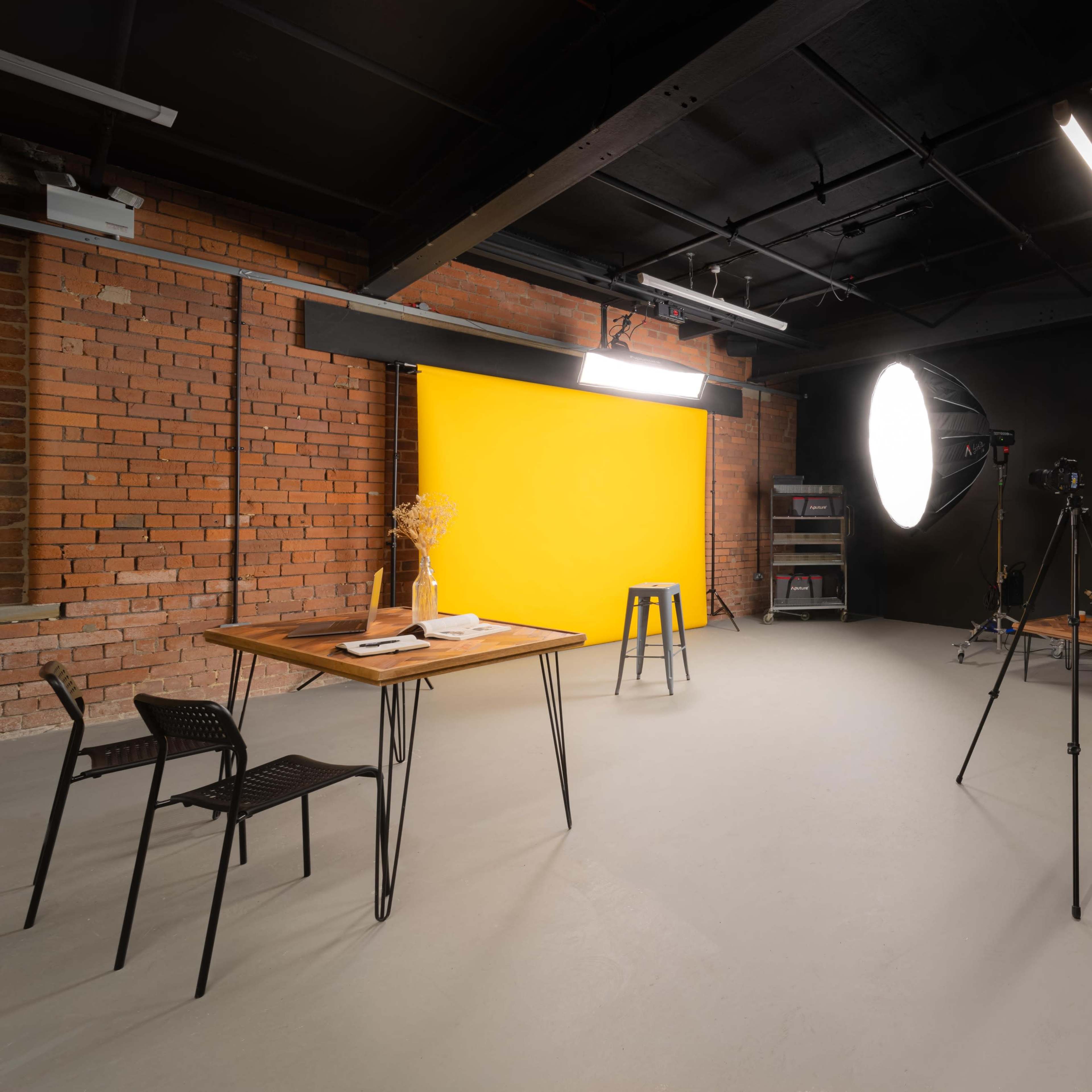 A photography studio featuring a yellow backdrop, lighting equipment, a camera on a tripod, a wooden table with a few items, and black chairs against exposed brick walls.