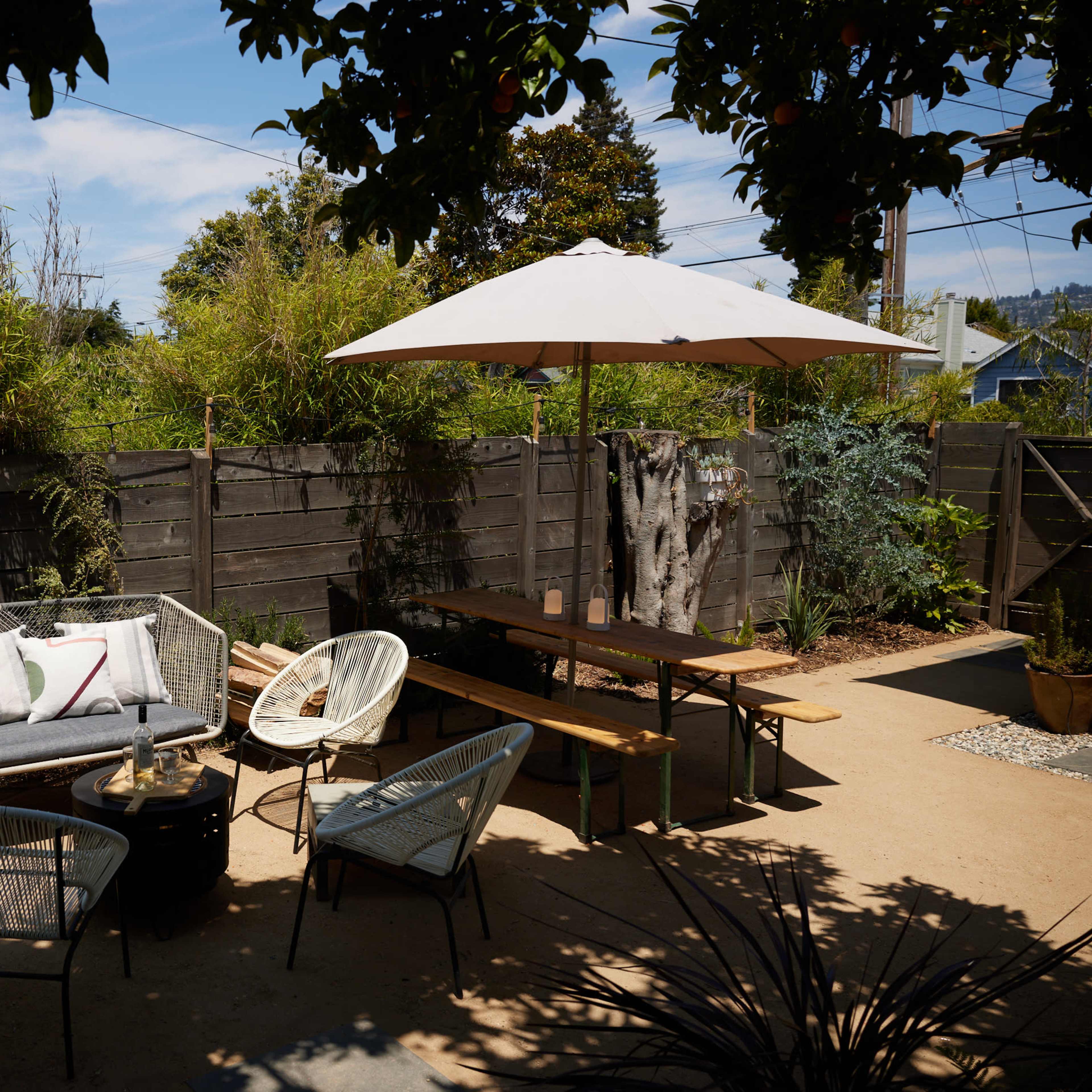 An outdoor patio area with a sofa, chairs, a table, and an umbrella, surrounded by plants and a wooden fence.