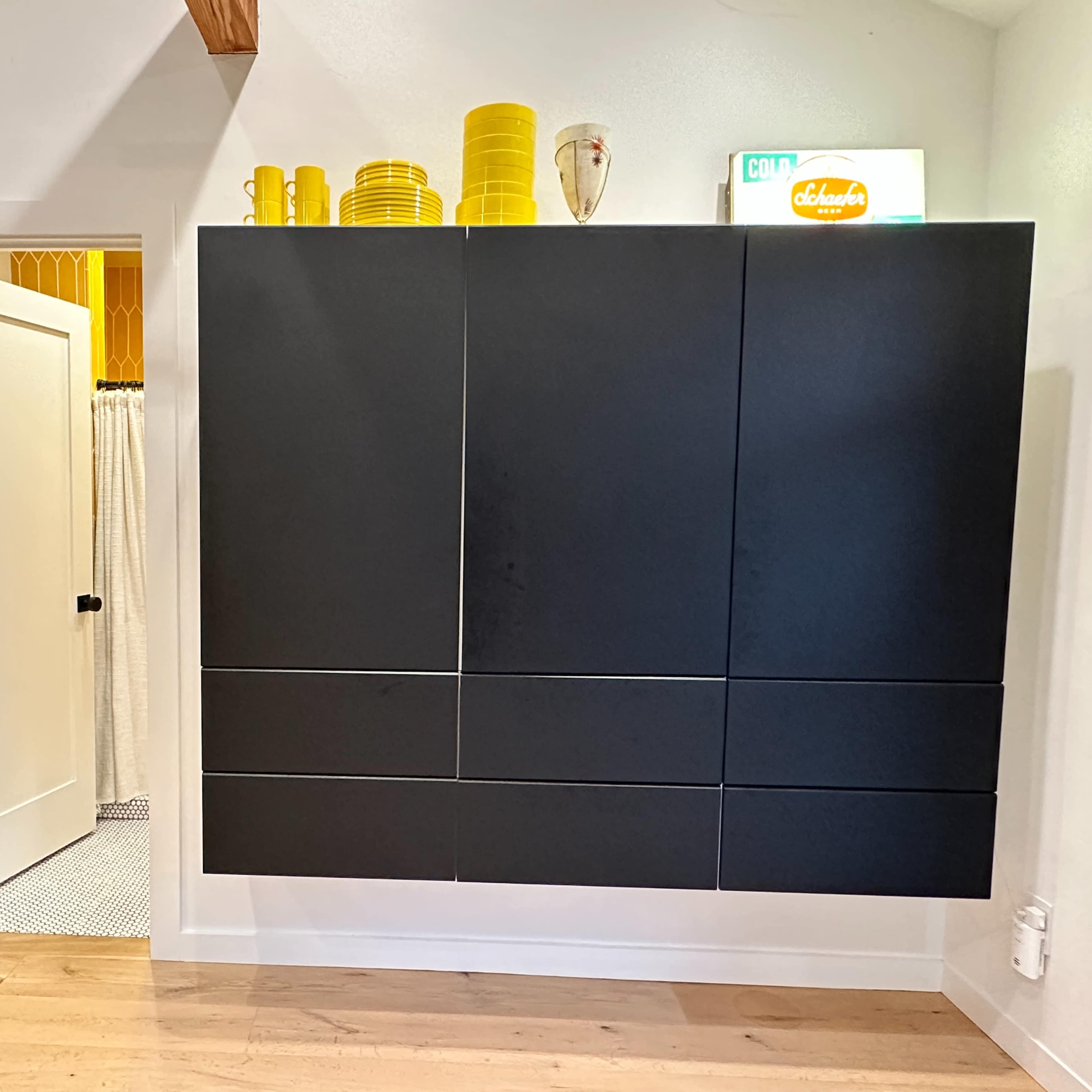 A contemporary kitchen area with black cabinetry, yellow dishware stacked on top, and a doorway leading to a bathroom.