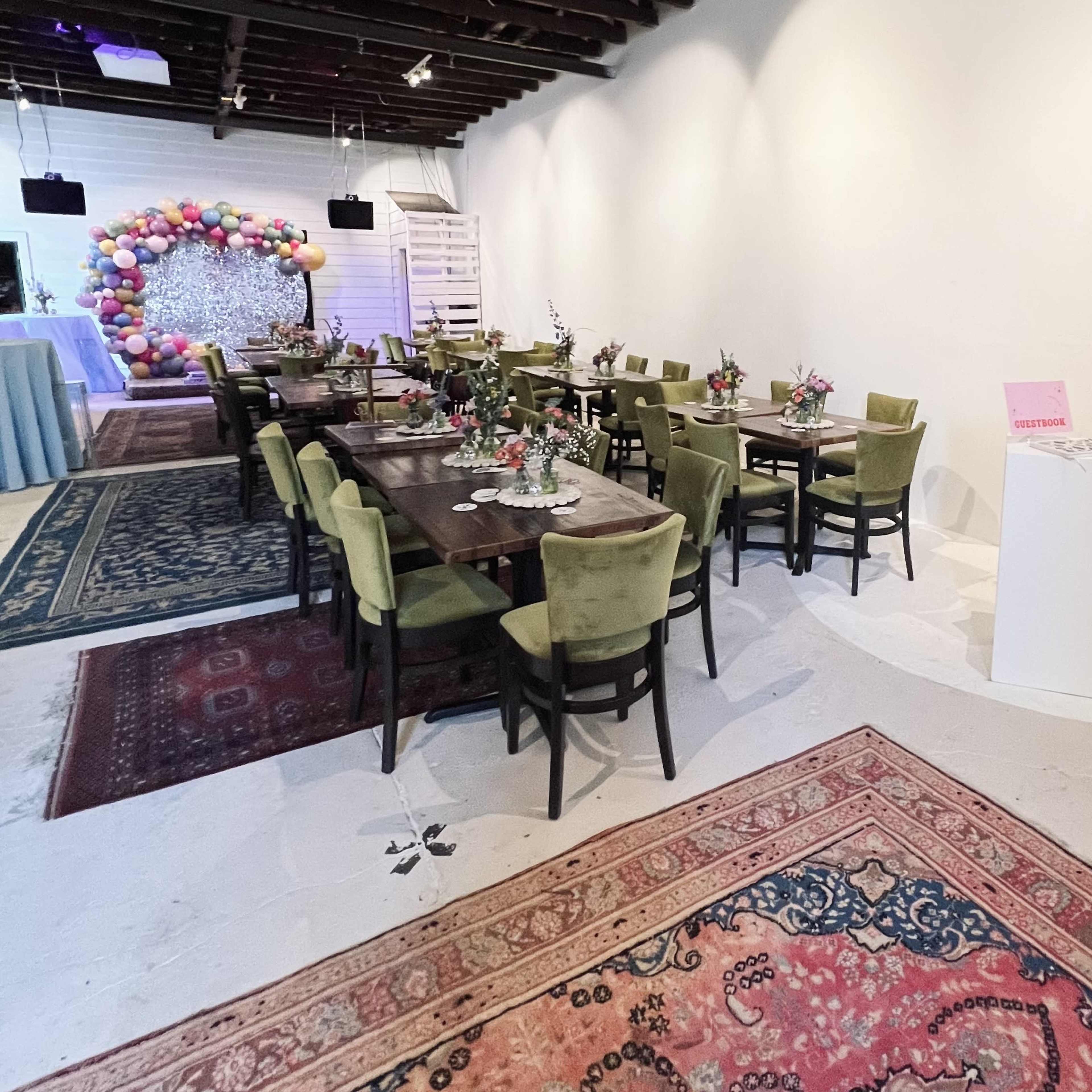 A spacious event room with several dining tables surrounded by green chairs, decorative floral arrangements, and a backdrop of colorful balloons.