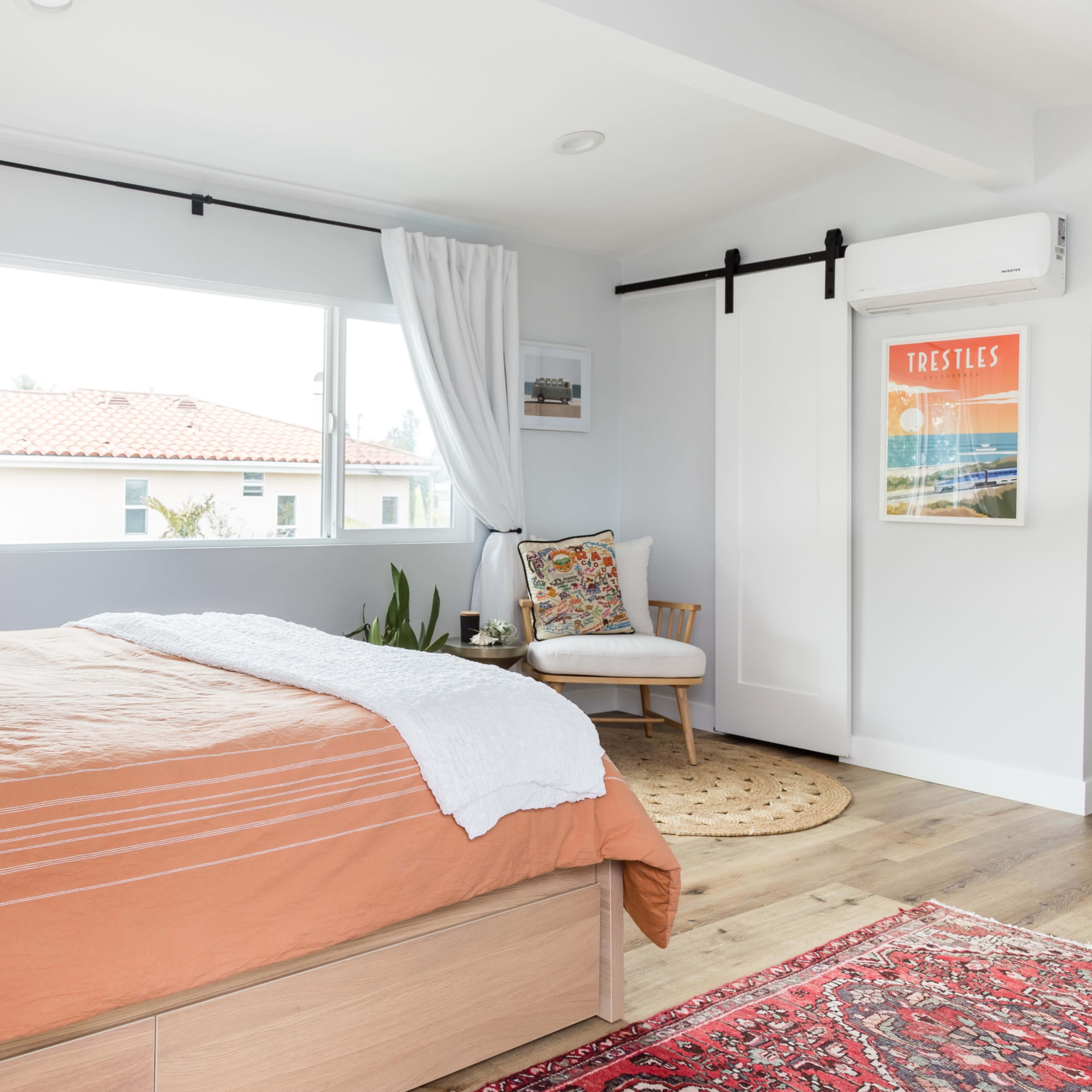 A bright bedroom with a bed covered in an orange duvet, a small decorative chair, and large windows allowing natural light to fill the space.