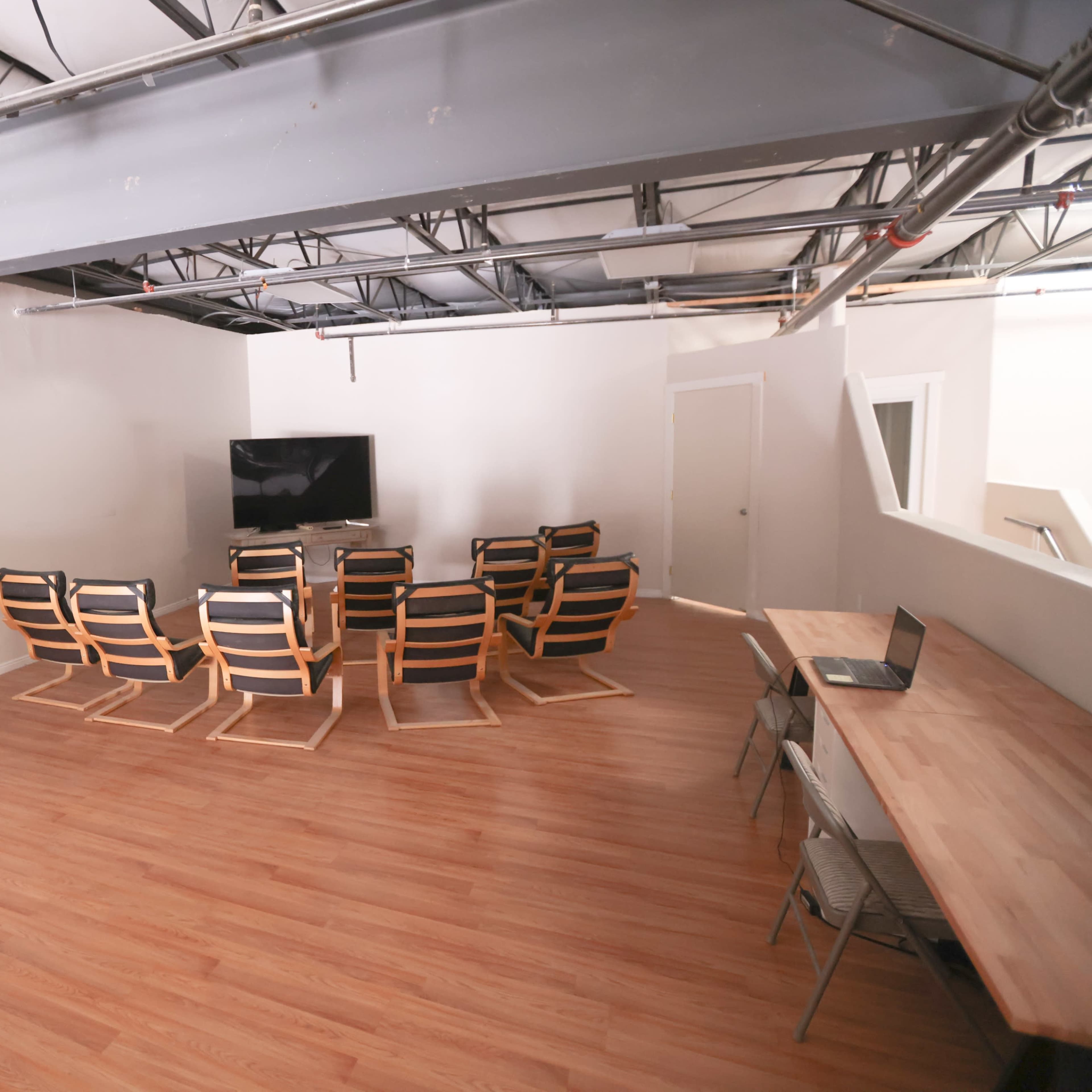 A spacious room with wooden flooring, featuring a seating arrangement of black and wooden chairs facing a television and a long table with chairs.