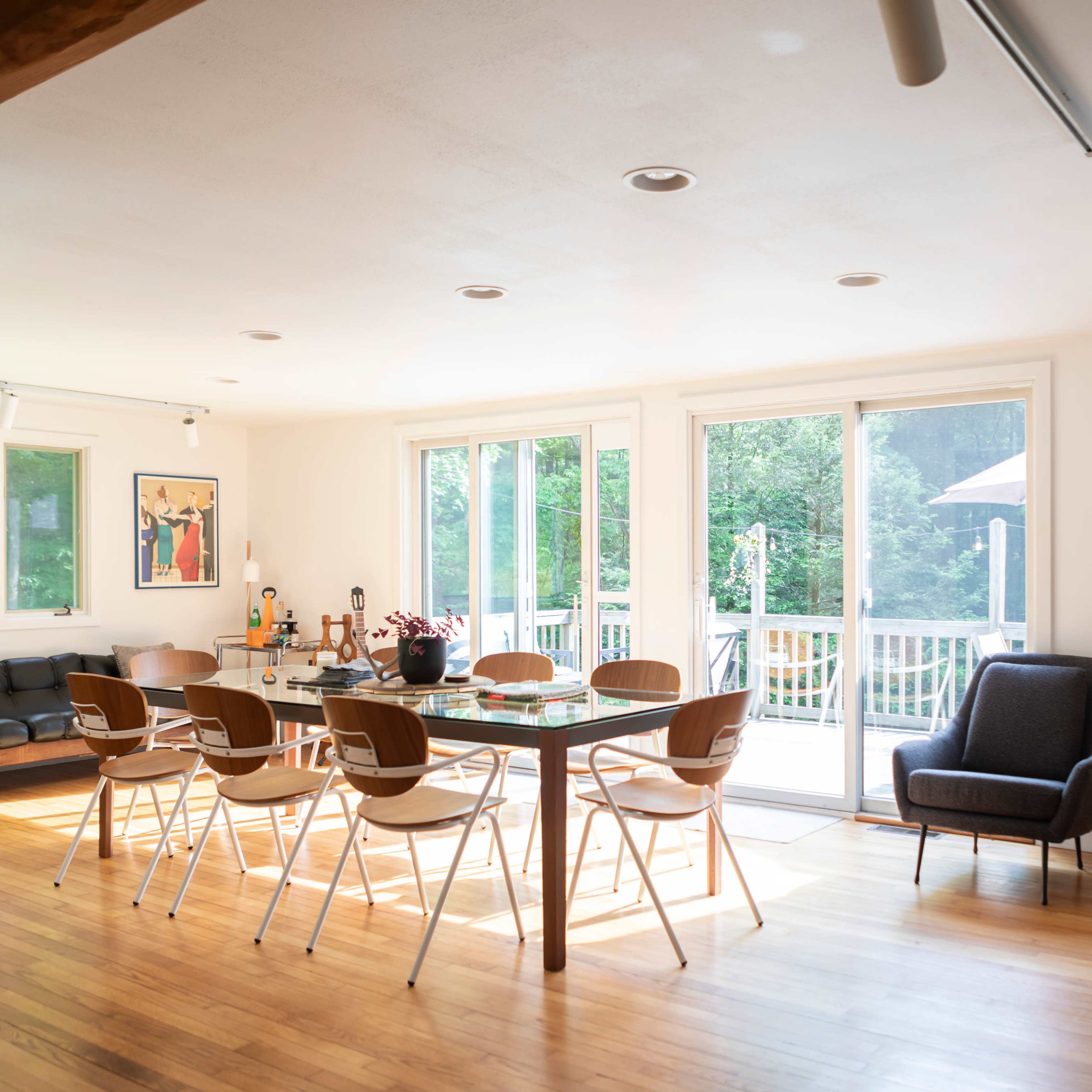 A bright, open living space featuring a dining area with a long table surrounded by chairs, a comfortable seating area, and large sliding glass doors leading to an outdoor deck.