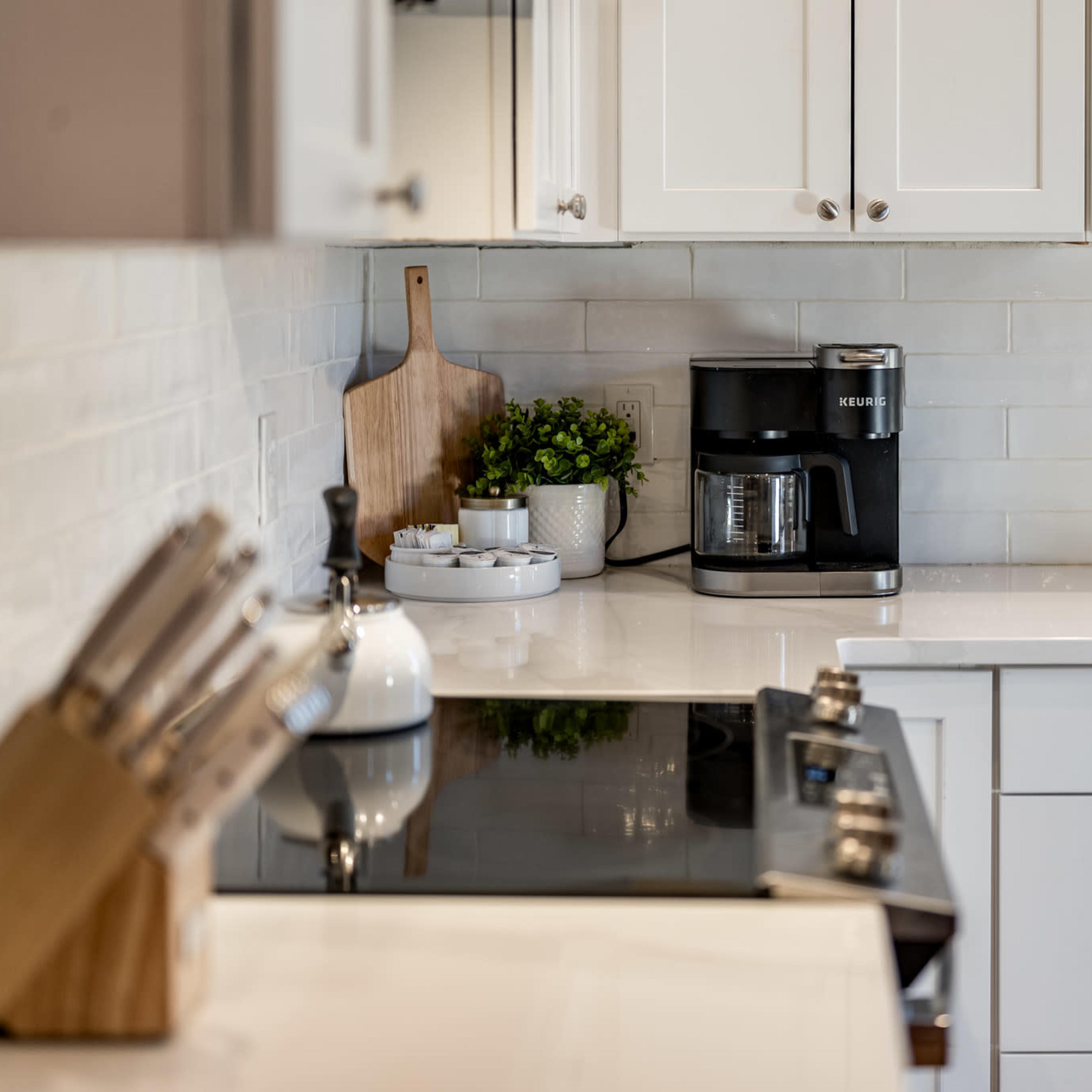 A modern kitchen features a black coffee maker on a countertop with white cabinetry and a wooden table.