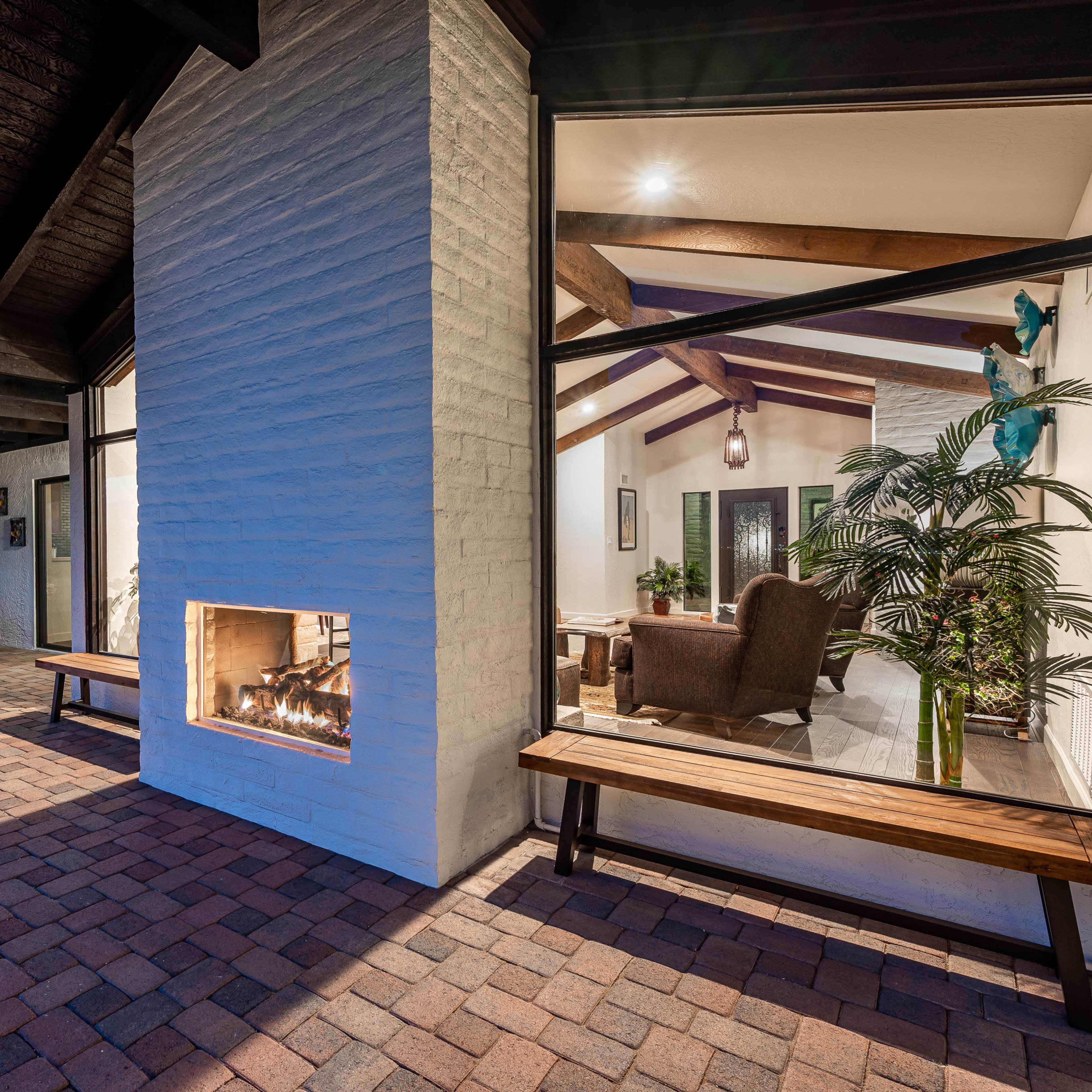 A modern home with an open patio area, large windows, and a cozy bench next to an outdoor fireplace, showcasing a view of the interior living space.