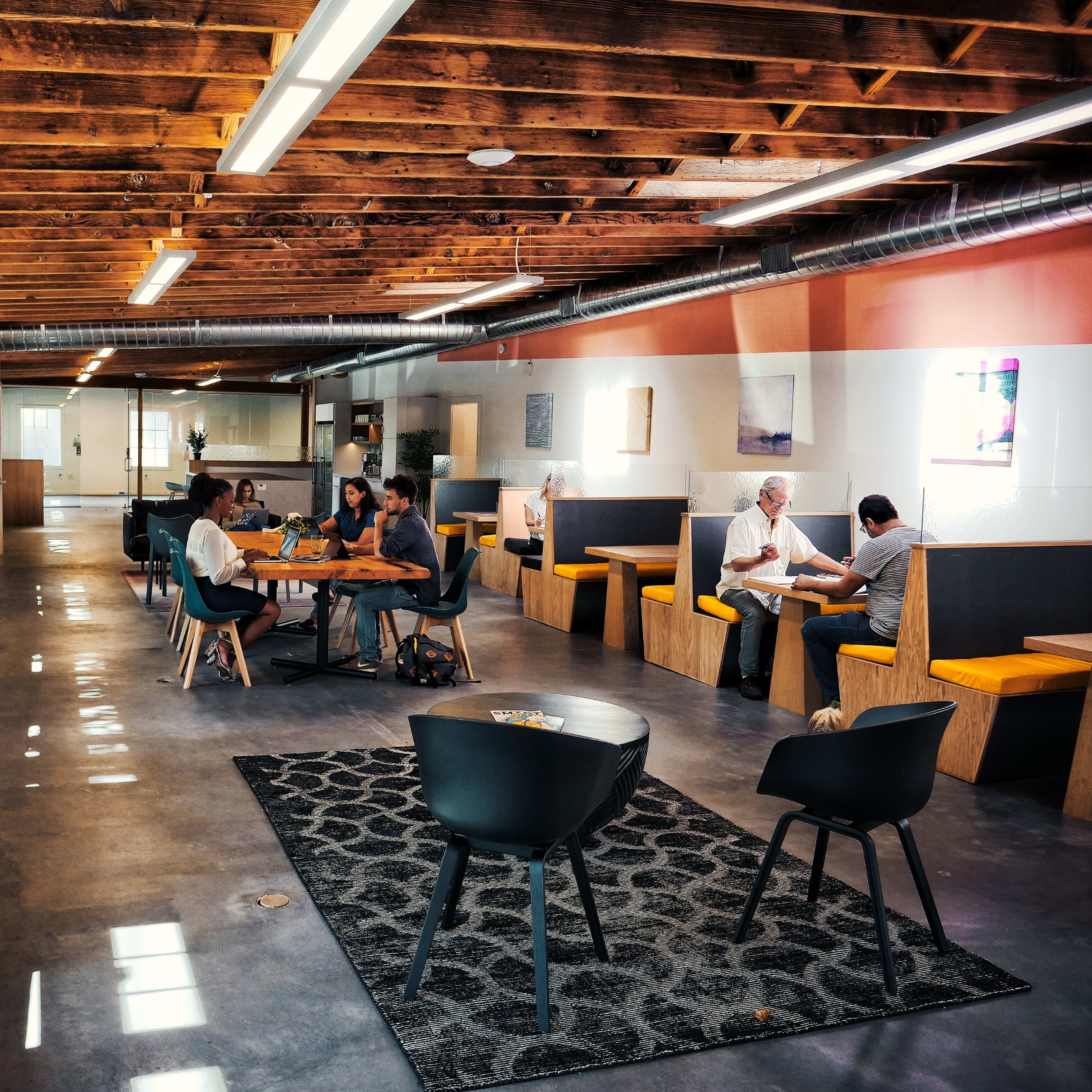 A modern open office space with wooden beams, various seating areas, and people engaged in work and conversation.