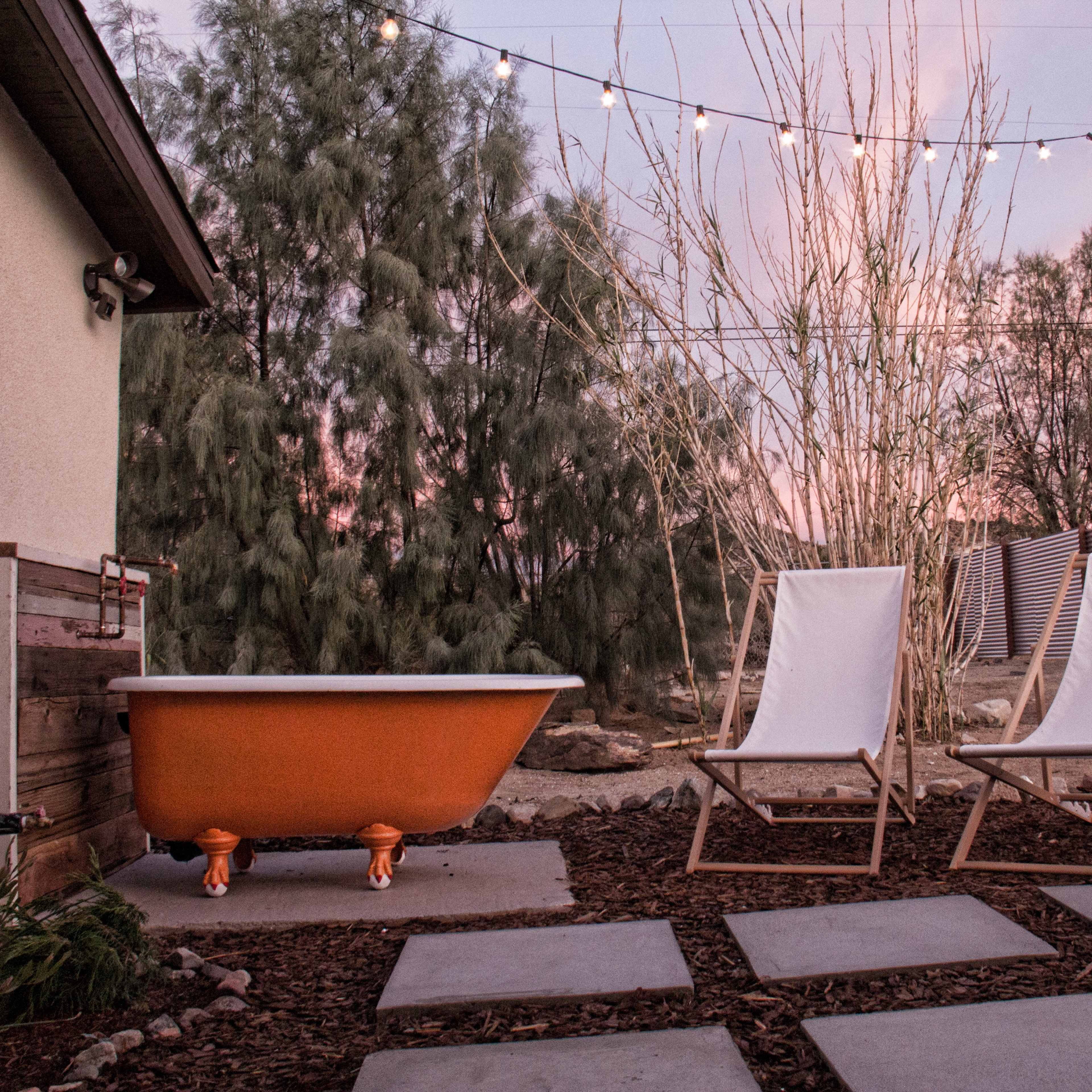 A backyard space featuring an orange clawfoot bathtub beside two white lawn chairs, with string lights overhead and trees in the background.